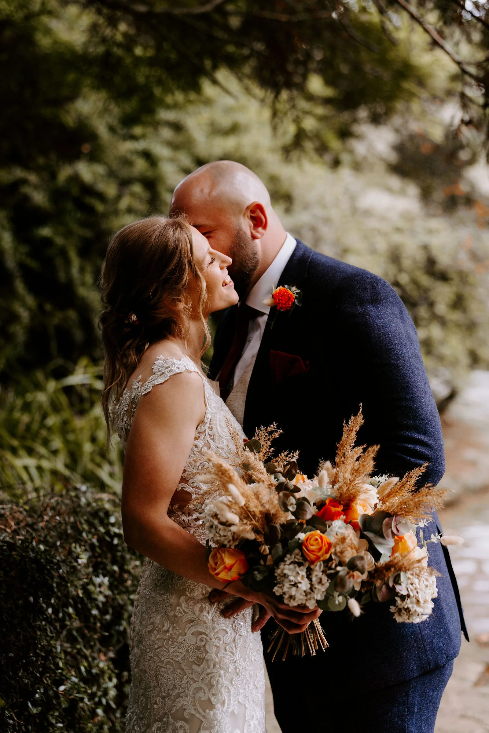 A bride in a lace gown smiles as her groom in a navy suit kisses her cheek, she holds a bouquet of dried grasses and orange roses, in dappled green shade