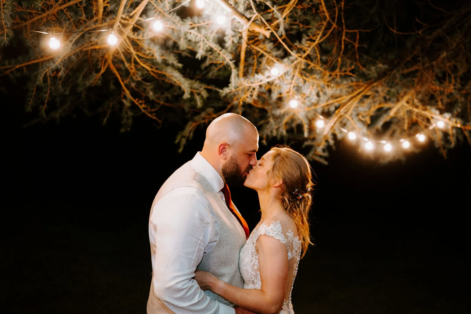 A groom in a pale waistcoat and a bride in a lace dress kiss at night beneath a tree hung with festoon lights