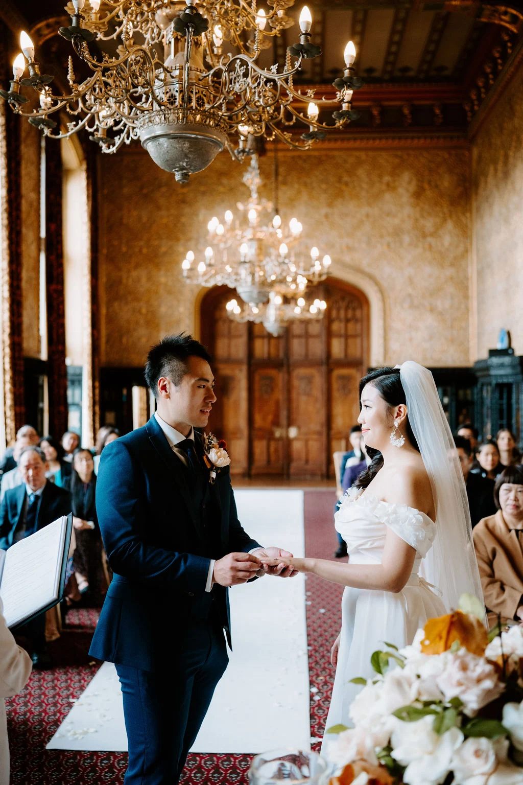 Yukki and Leslie holding hands during their ceremony beneath chandeliers at Carlton Towers
