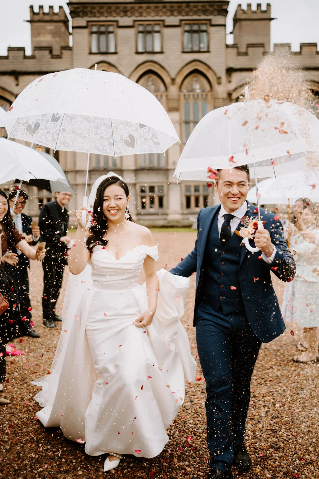 Yukki and Leslie walking through confetti beneath clear umbrellas at Carlton Towers