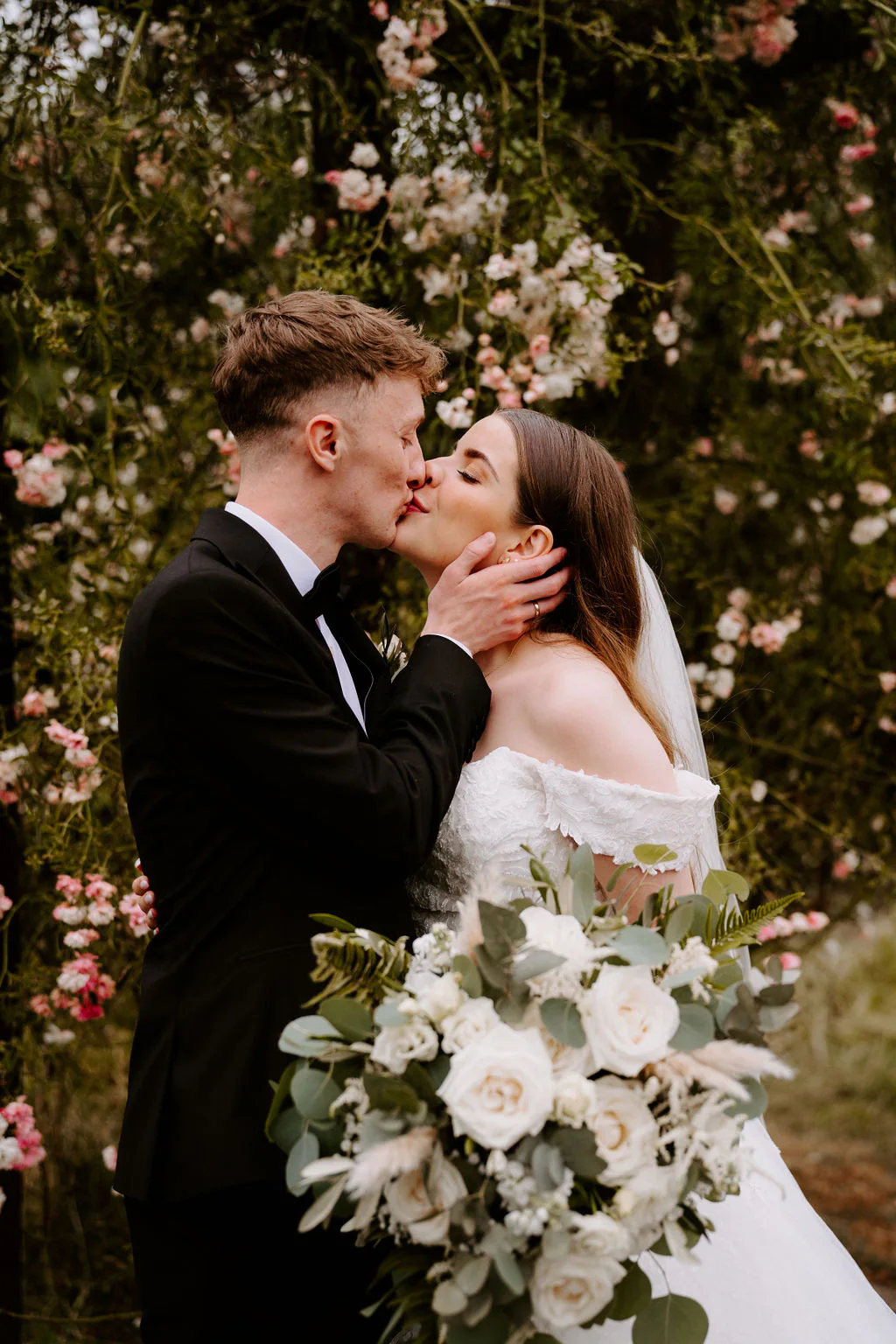 Noomi and Jamie kiss among pink roses and blossom, Noomi holding a bouquet of white flowers