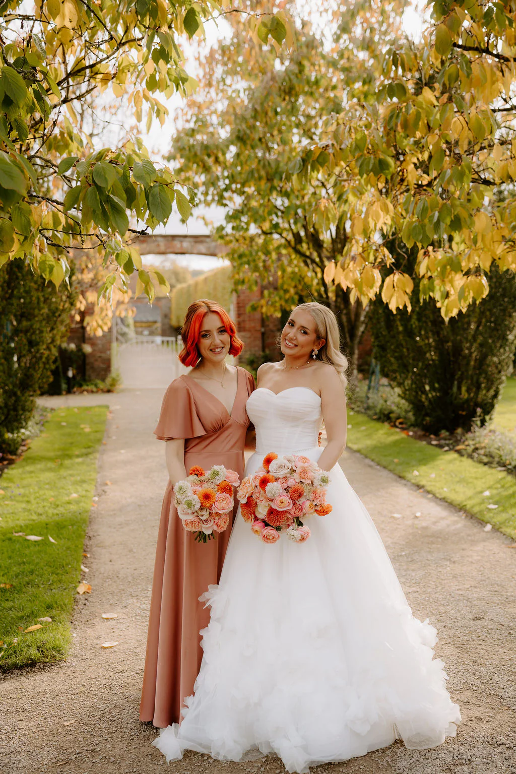 A bride and a bridesmaid in a dusty-pink satin dress hold orange and pink bouquets on a gravel path under yellowing leaves, with a brick archway and white gate behind them
