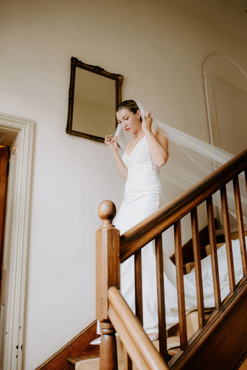 A bride in a white gown with a veil held to one side comes down a wooden staircase with a polished newel post beside a gilt mirror on the wall