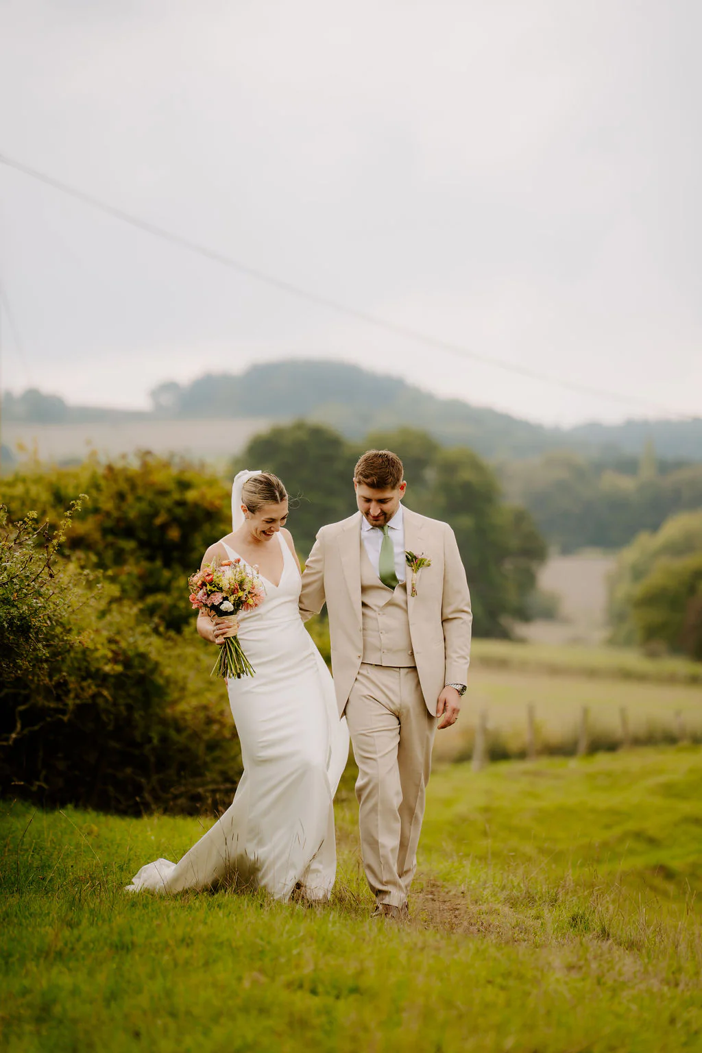 A bride in a fitted white gown holding a bright bouquet and a groom in a cream three-piece suit with a green tie walk arm in arm across a green field with hedges and wooded hills behind them