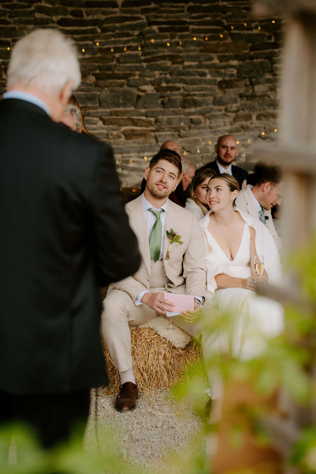 A groom in a cream suit and a bride in white sit smiling on hay bales in front of a slate stone wall hung with fairy lights while a man in a dark jacket speaks in the foreground