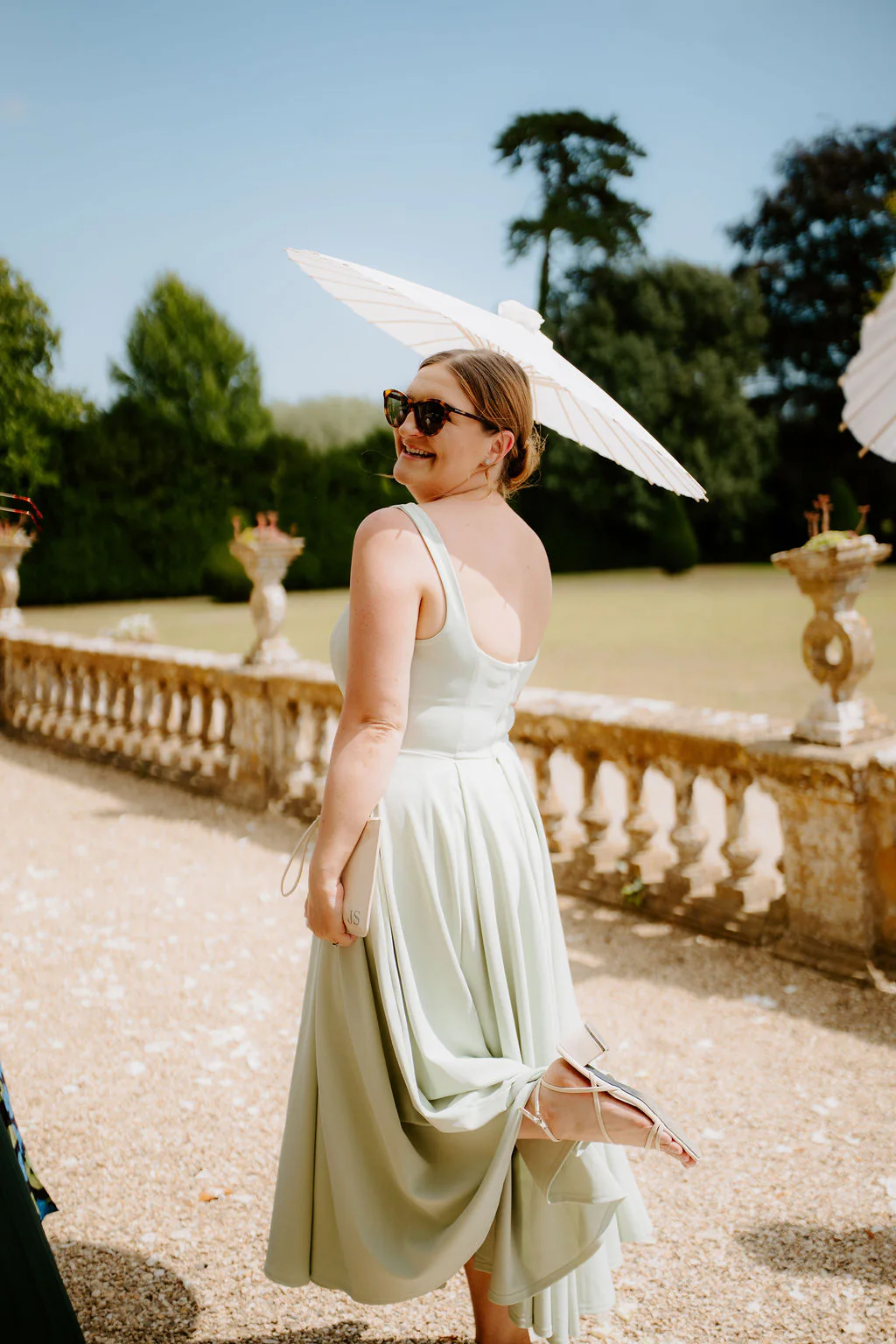 A guest in a sage-green dress looks back over her shoulder holding a white parasol in front of a stone balustrade at Brympton House