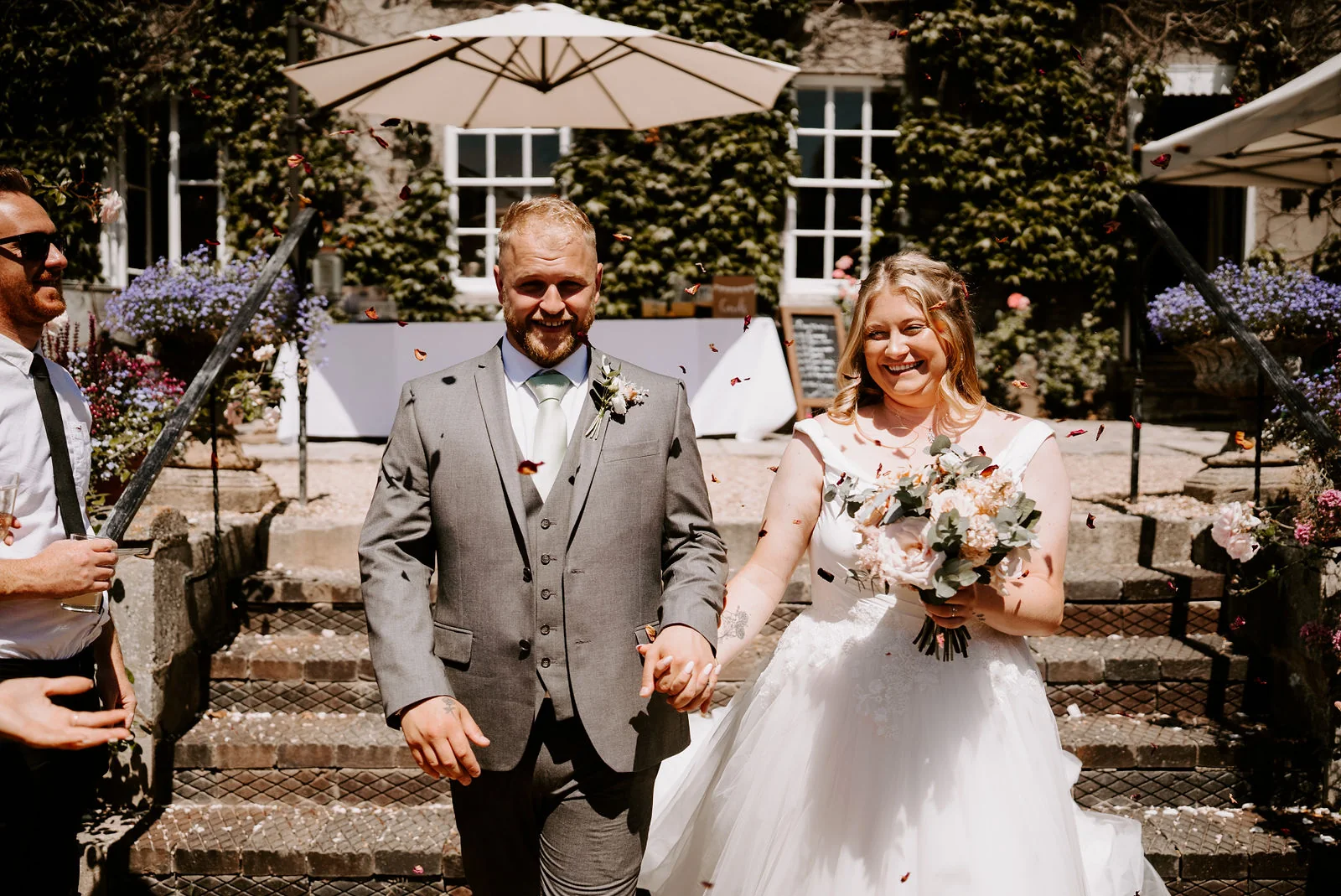 A groom in a grey suit and his bride holding a bouquet walk down stone steps towards the camera, smiling, with an ivy-covered house and a white umbrella behind them at Pennard House