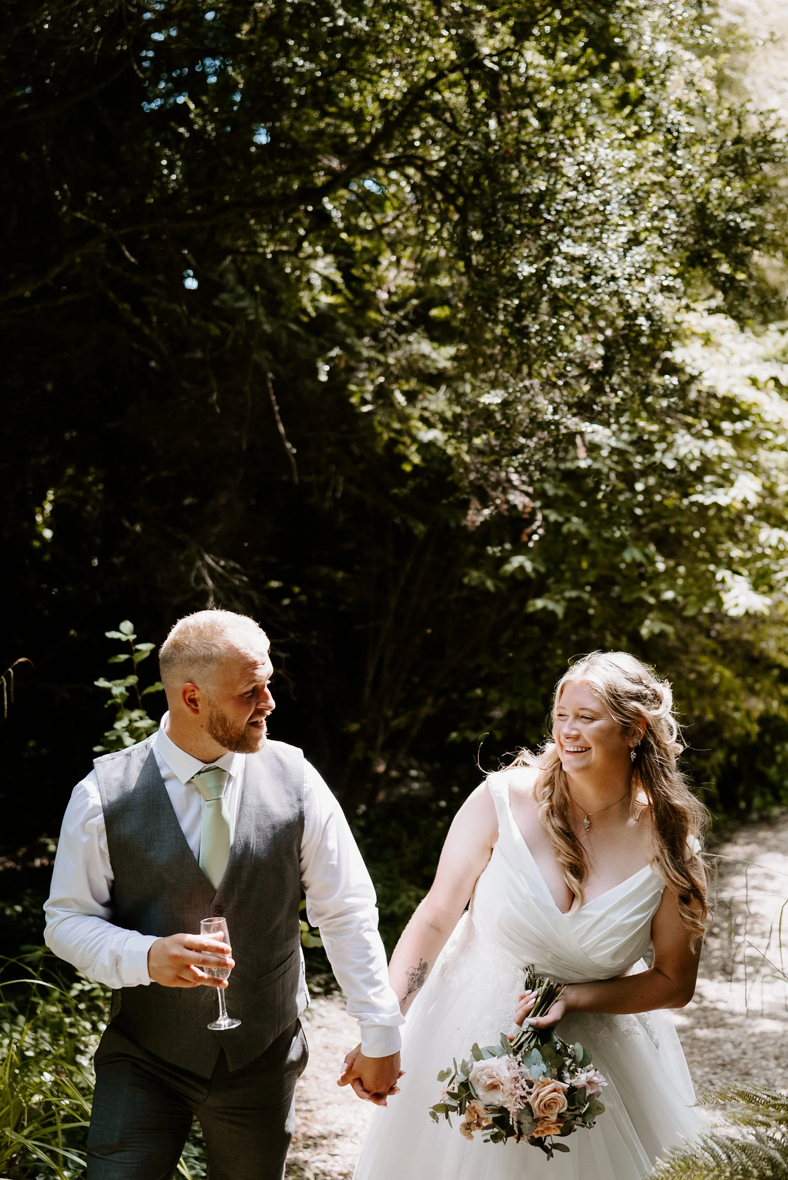 A groom in a waistcoat holding a glass walks hand in hand with his laughing bride, who carries a bouquet, through woodland at Pennard House