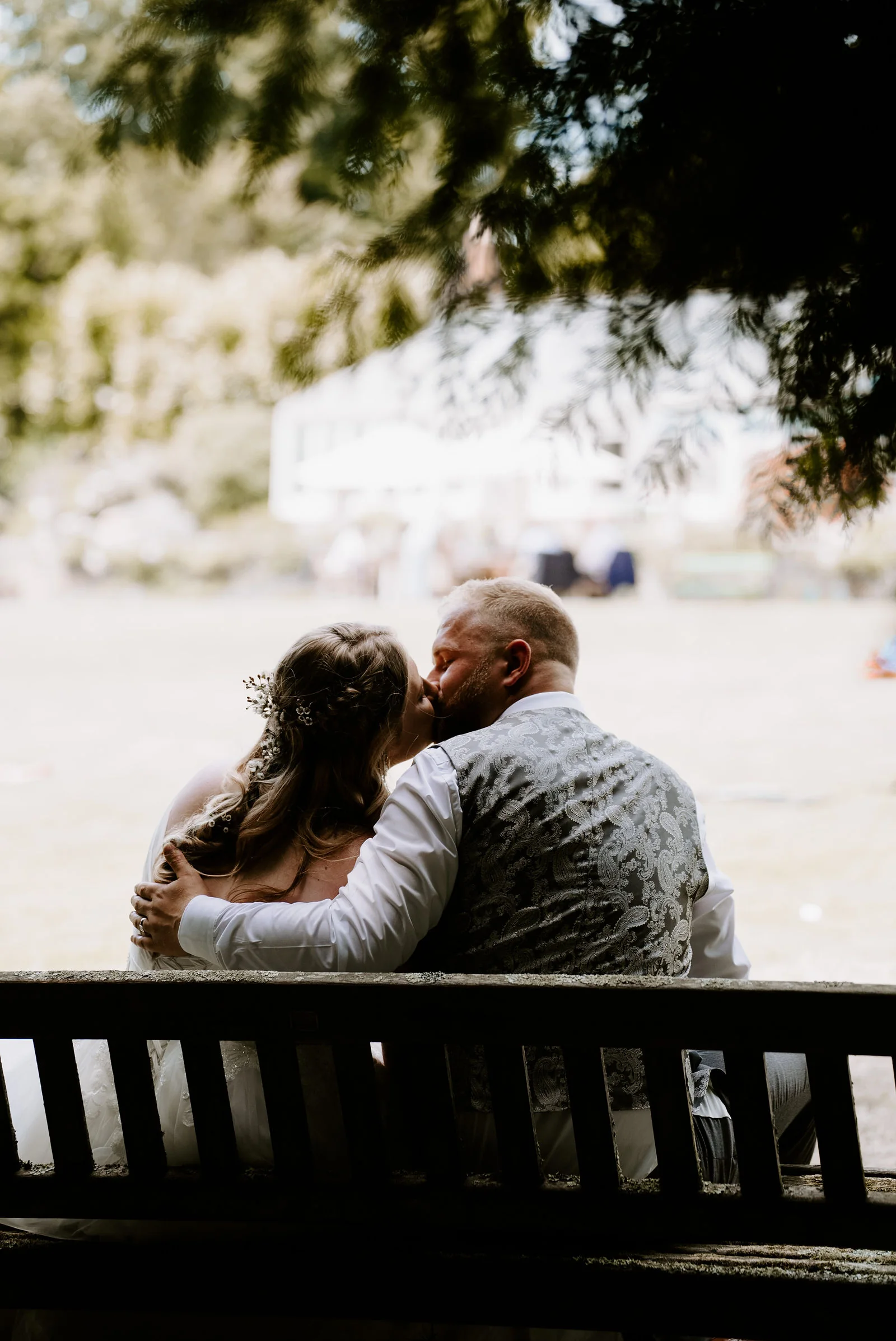 A bride and groom kiss as they sit close together on a wooden bench, seen from behind, looking across a lawn towards a distant white building at Pennard House