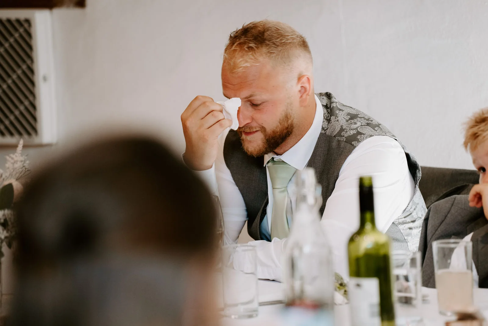 A bearded groom in a waistcoat wipes his eye with his hand during the speeches at the wedding breakfast at Pennard House