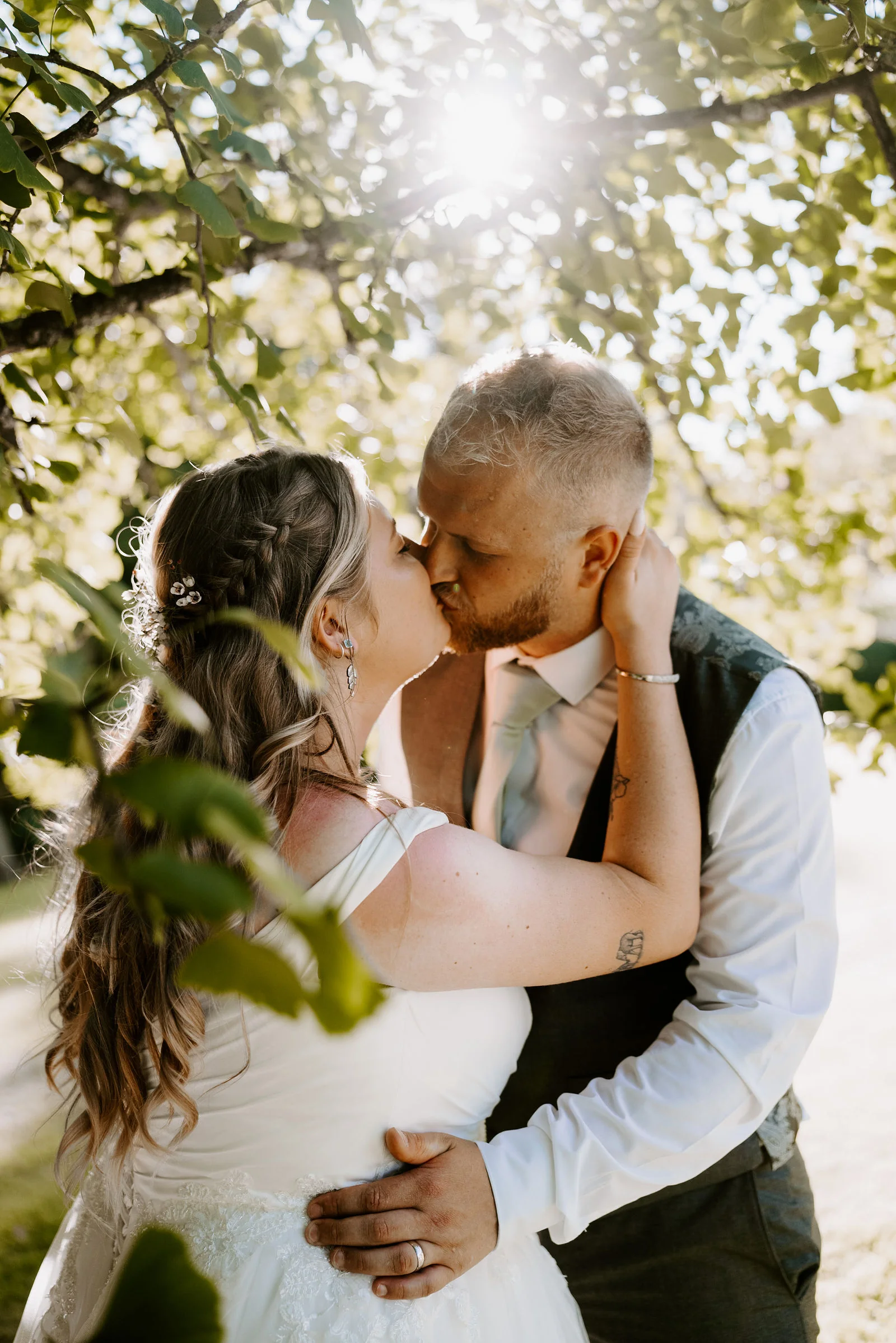 A groom in a waistcoat kisses his bride under leafy branches with the low sun flaring through them at Pennard House