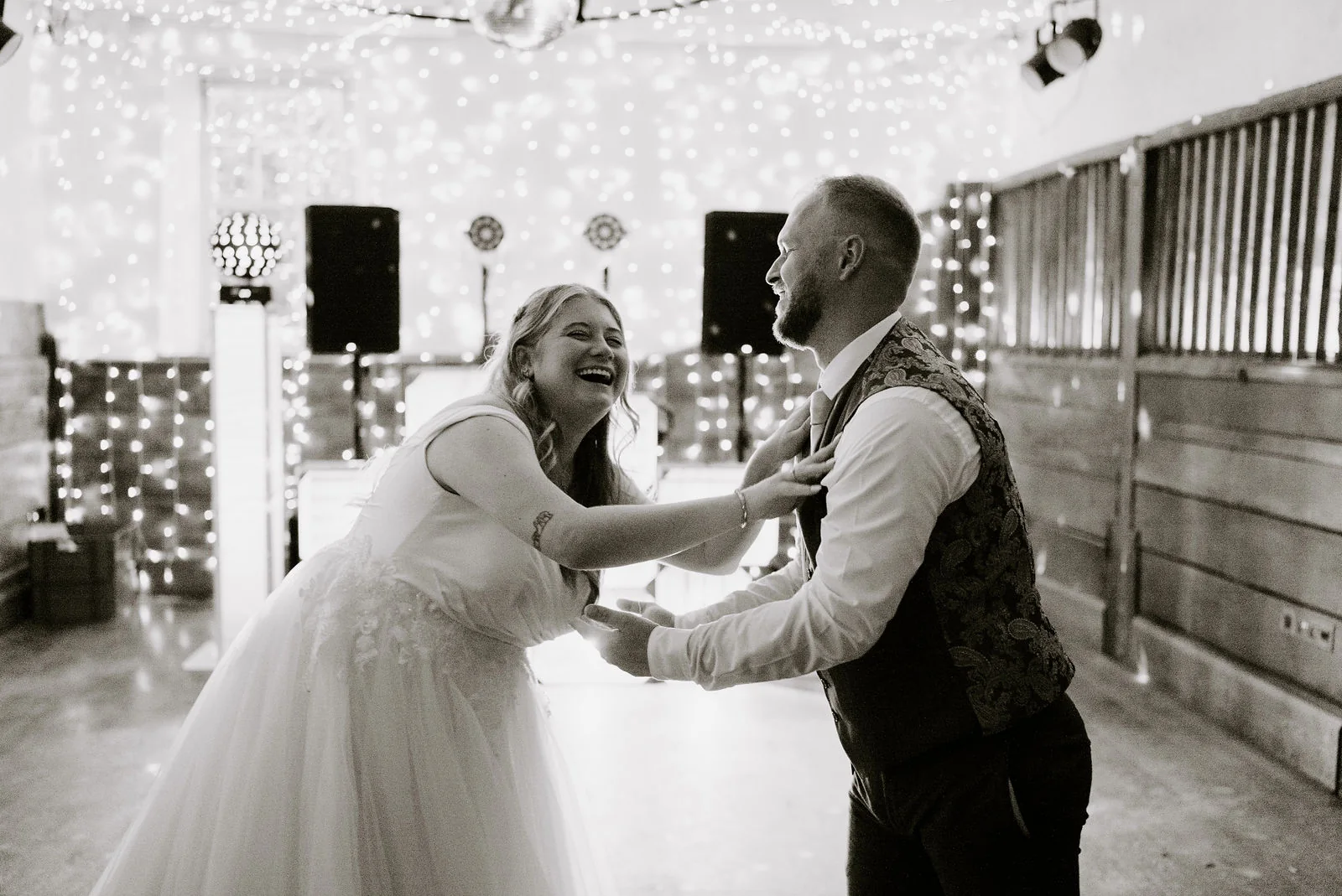 In black and white, a bride laughs as she dances with her groom, their hands joined, in a room hung with string lights at Pennard House