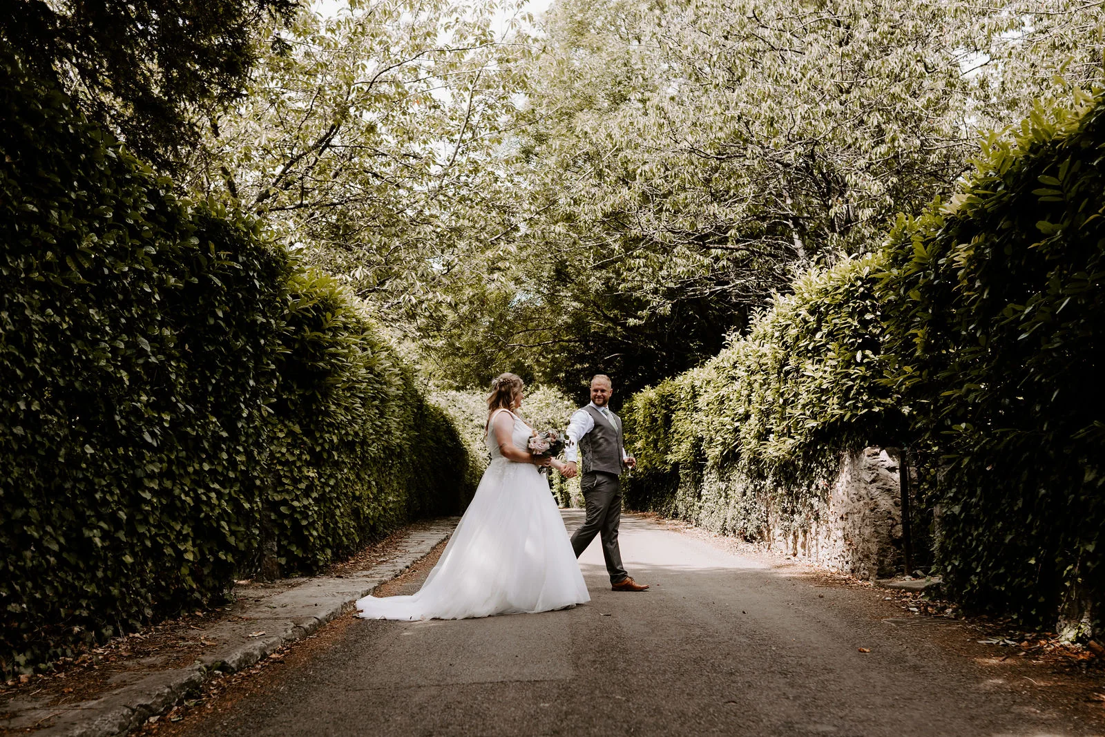 A bride in a full-skirted gown and a groom stand together in the middle of a tree-lined lane between tall hedges near Pennard House