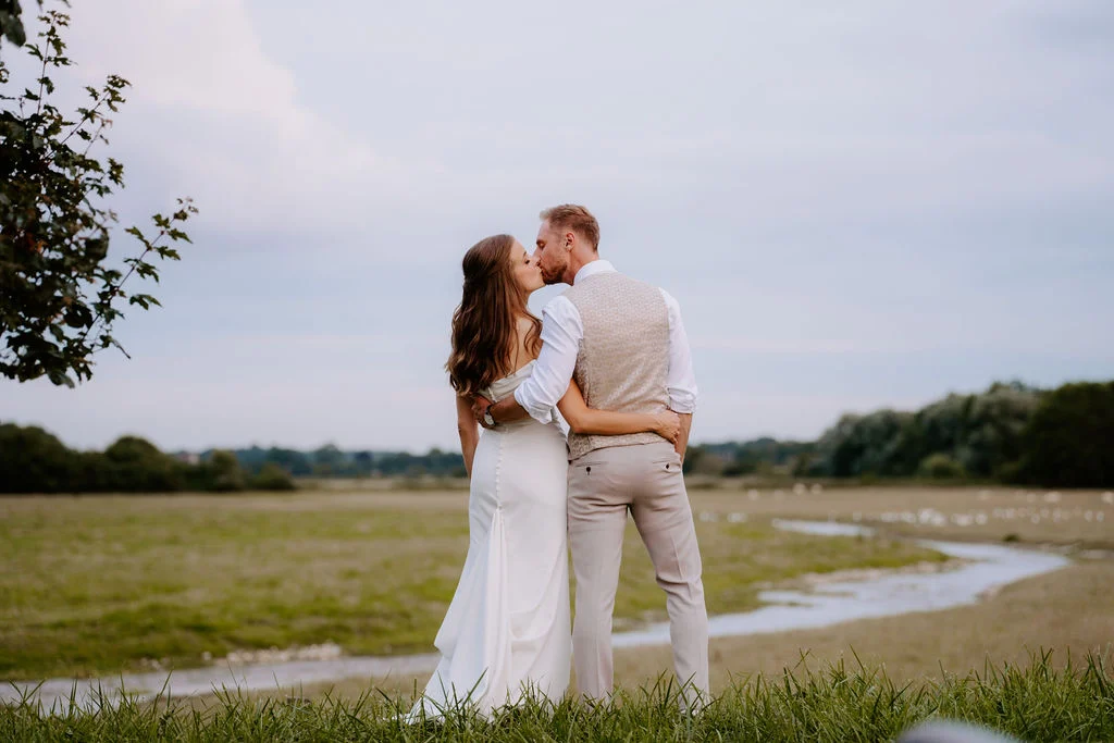 Hannah and Adam embracing beside a lake with countryside beyond