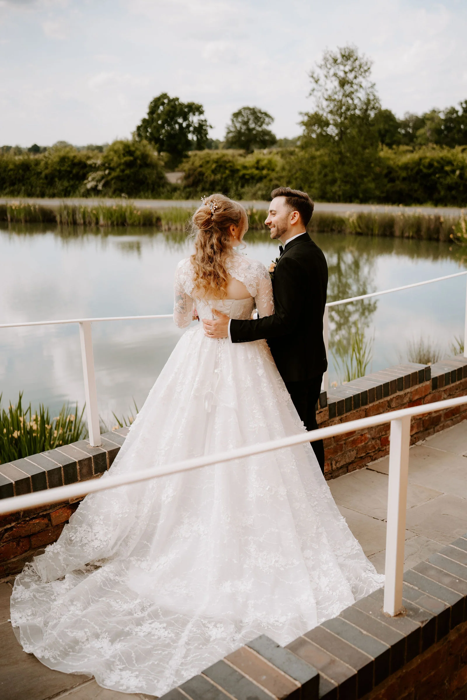 A bride in a lace-sleeved gown with a long train and a groom in black stand on a brick terrace with white railings above a lake