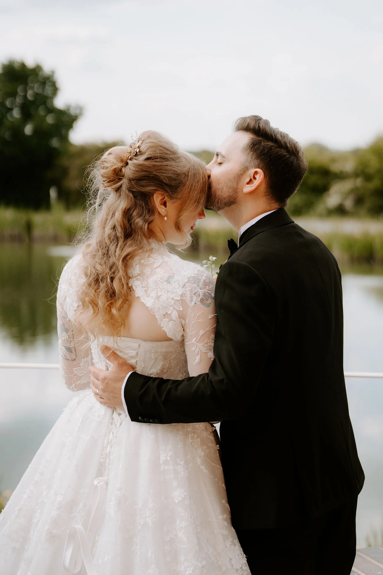 A groom in black tie kisses the forehead of a bride in a lace gown by a lake with reeds behind