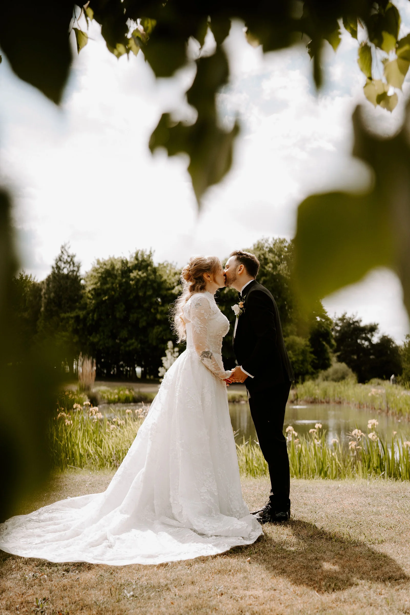A bride in a lace gown with a long train and a groom in black kiss on a lawn, framed by out-of-focus leaves, with a lake and iris beds behind