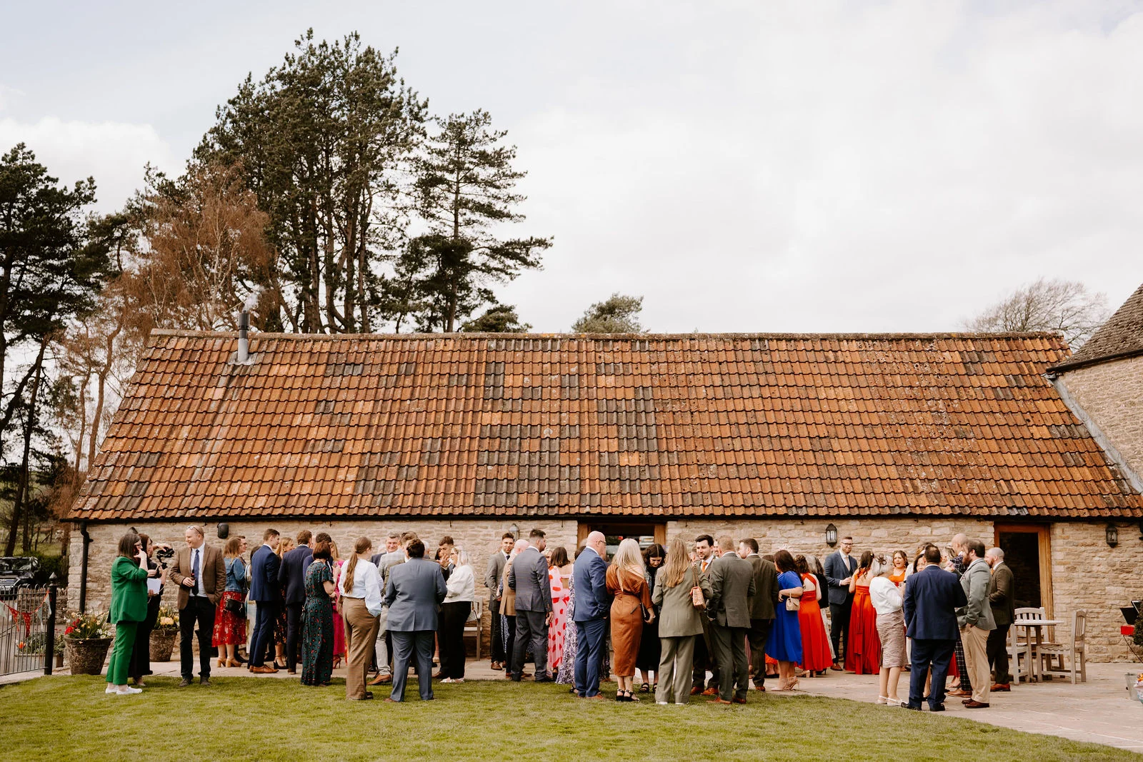 Wedding guests in smart outfits stand chatting in groups on the lawn and terrace in front of a long stone barn with an orange tiled roof, with tall pine trees rising behind it under a pale sky.