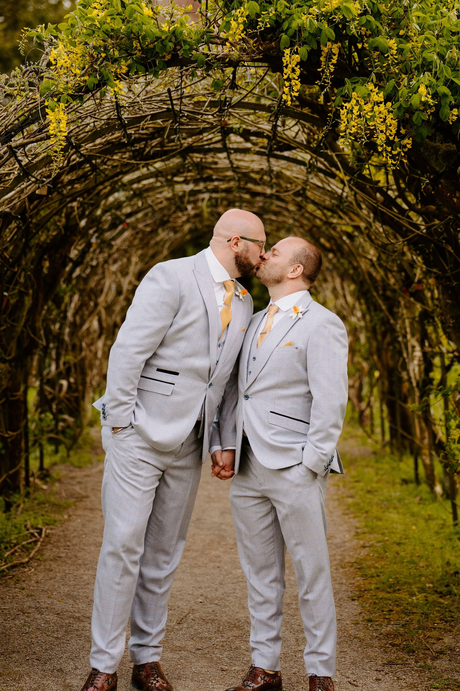 Two men in pale grey suits with yellow ties kiss and hold hands on a gravel path beneath a tunnel of woven branches hung with yellow blossom.