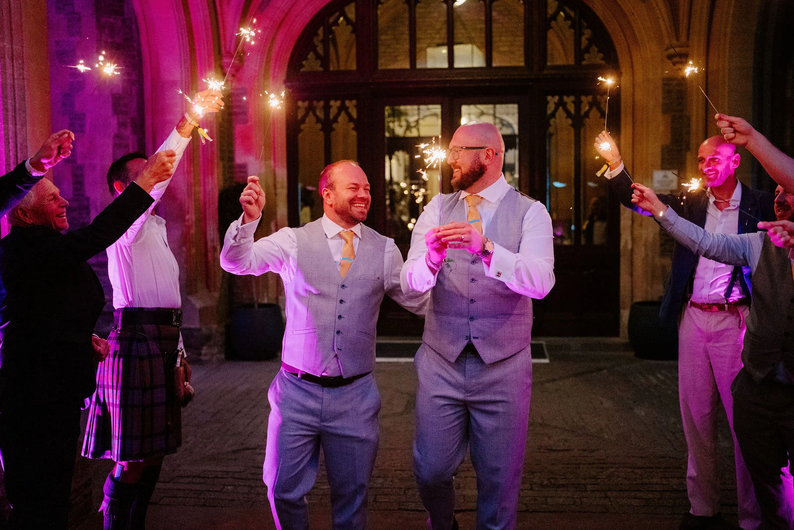 Two men in grey waistcoats and white shirts walk laughing between friends holding sparklers aloft, lit in pink in front of an arched stone doorway at night.