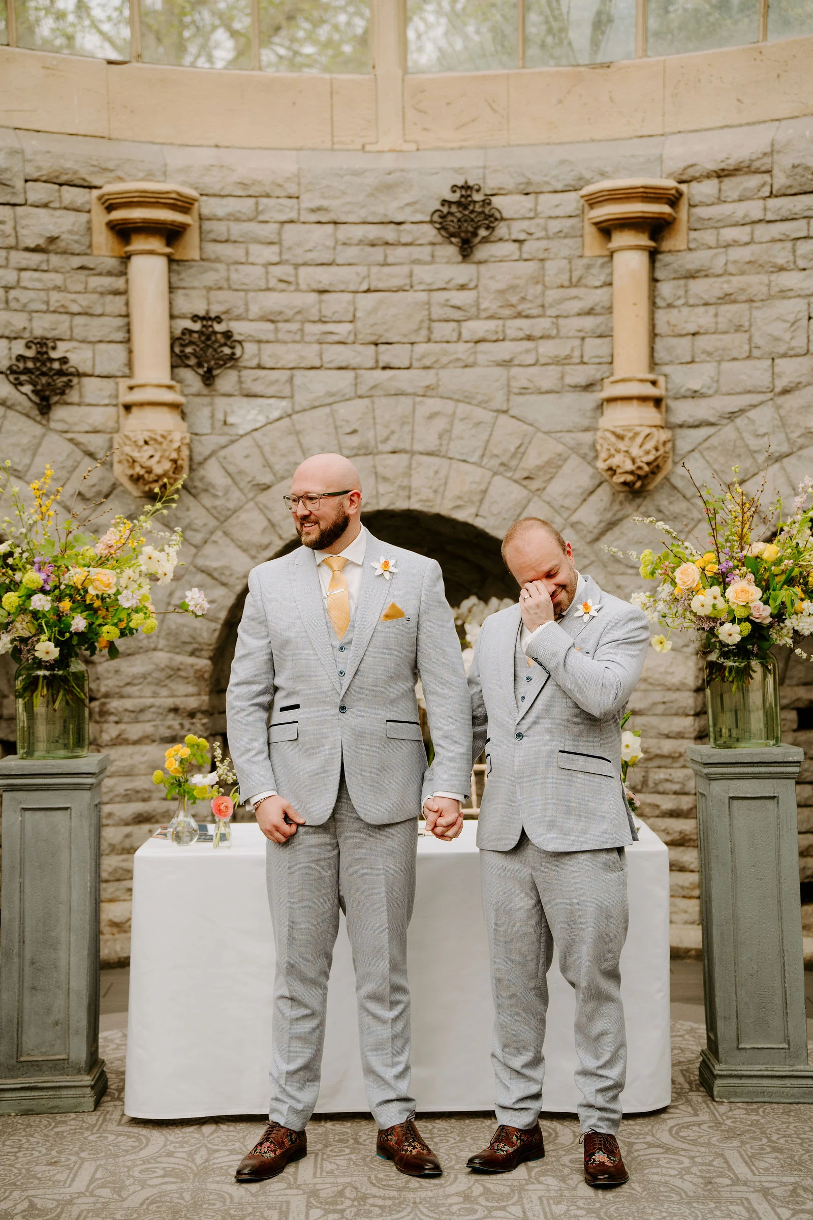 Two men in light grey three-piece suits with yellow ties stand hand in hand in front of a white draped table and spring flowers in a stone-walled round room, one looking to the side and the other wiping a tear from his eye.