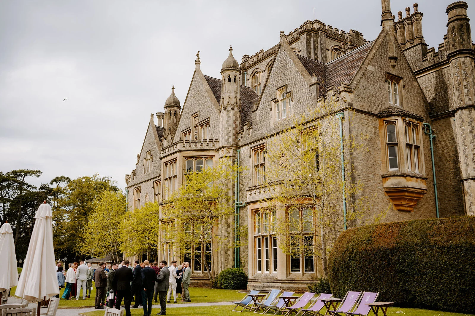 A large pale stone country house with pointed gables, tall chimneys and bay windows, seen from the lawn under a cloudy sky, with guests chatting in groups beside deckchairs and closed parasols.
