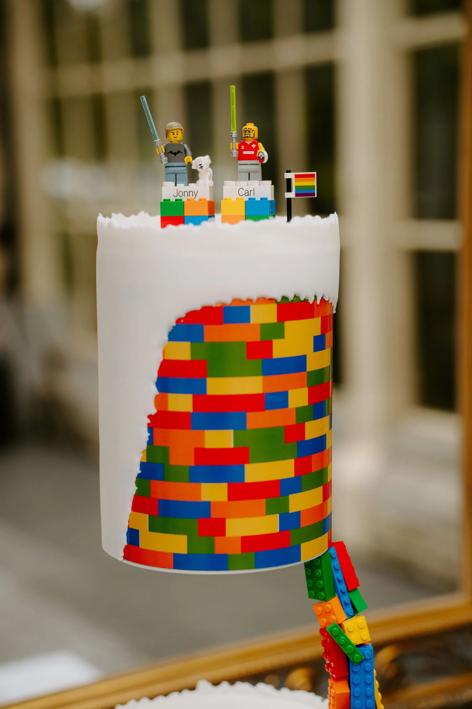 A tall white cake with a bright pattern of Lego bricks down one side, two small Lego figures and a rainbow flag on top, photographed in front of a tall window.
