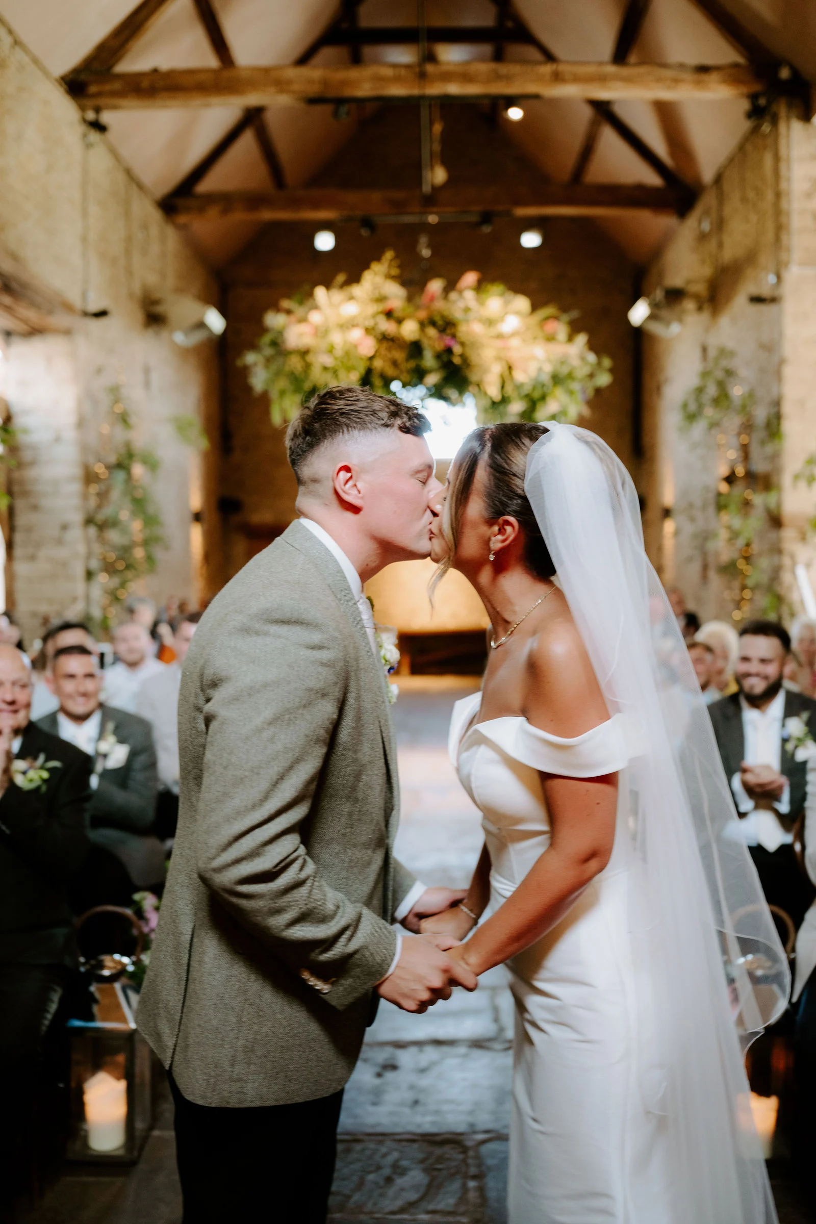 A groom in a green-grey tweed jacket and a bride in an off-the-shoulder gown and long veil kiss while holding hands in a stone barn, with smiling seated guests and a hanging floral installation behind them.