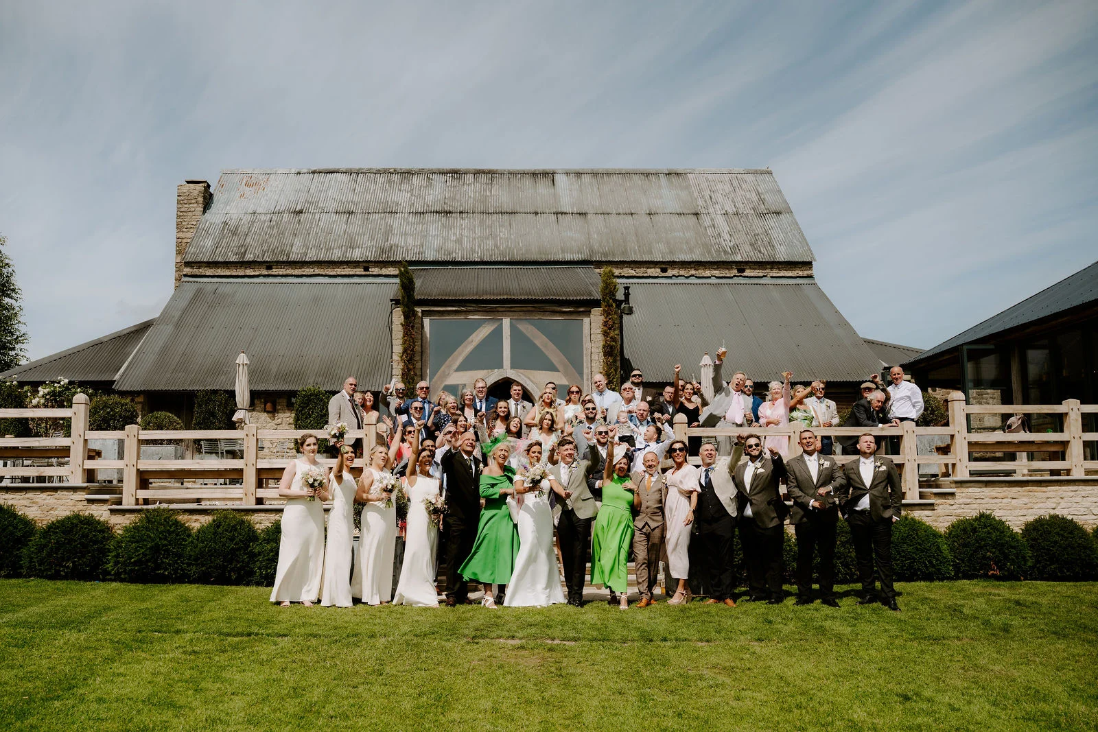 A large wedding party stands on a lawn with arms raised and cheering, in front of a long stone barn with a grey corrugated roof, a tall glazed gable and a timber-railed terrace, under a pale blue sky.
