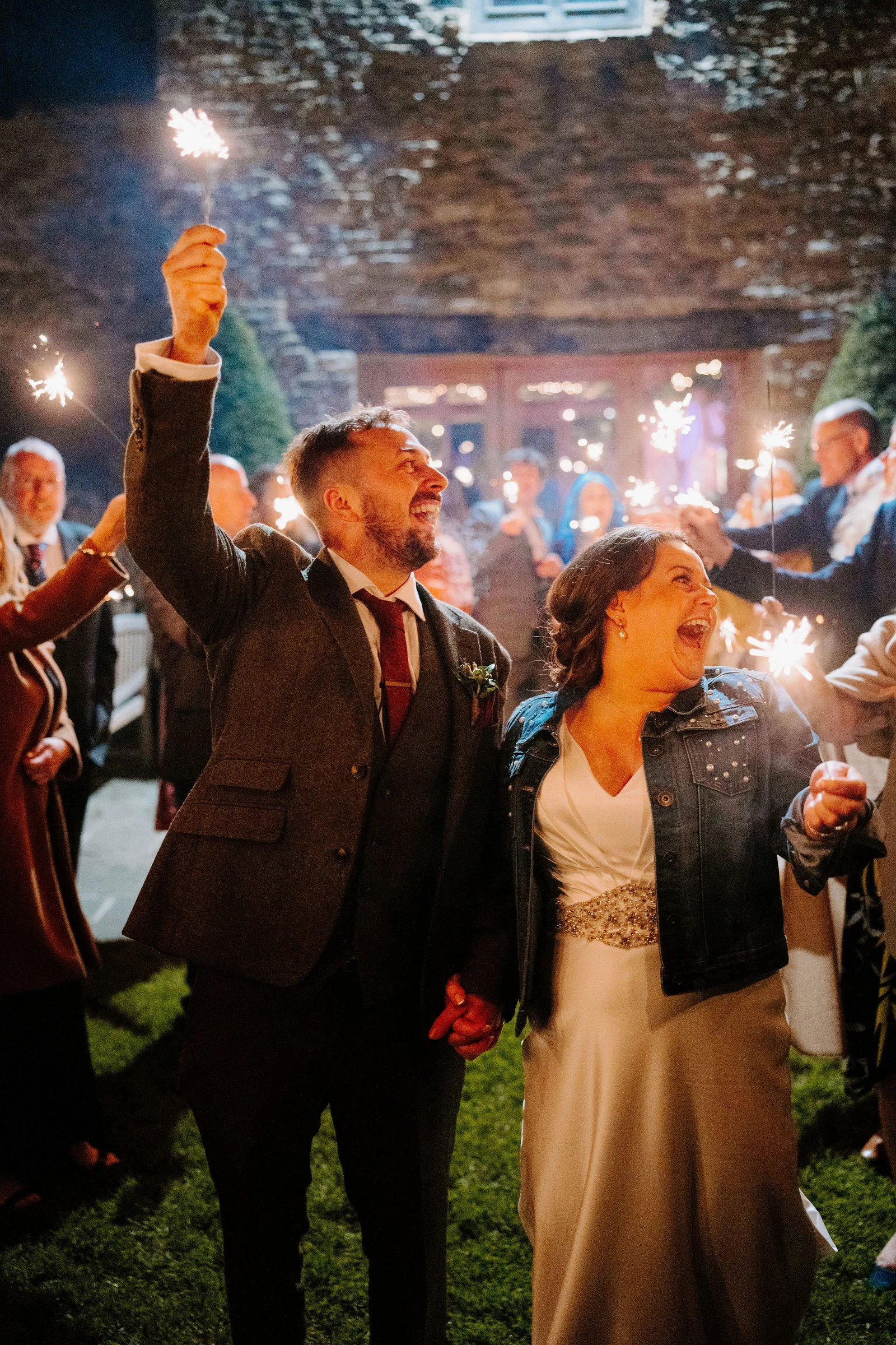 A groom raises a lit sparkler above his head while his laughing bride, in a denim jacket over her wedding dress, holds his hand, with guests holding sparklers behind them on the lawn at night.