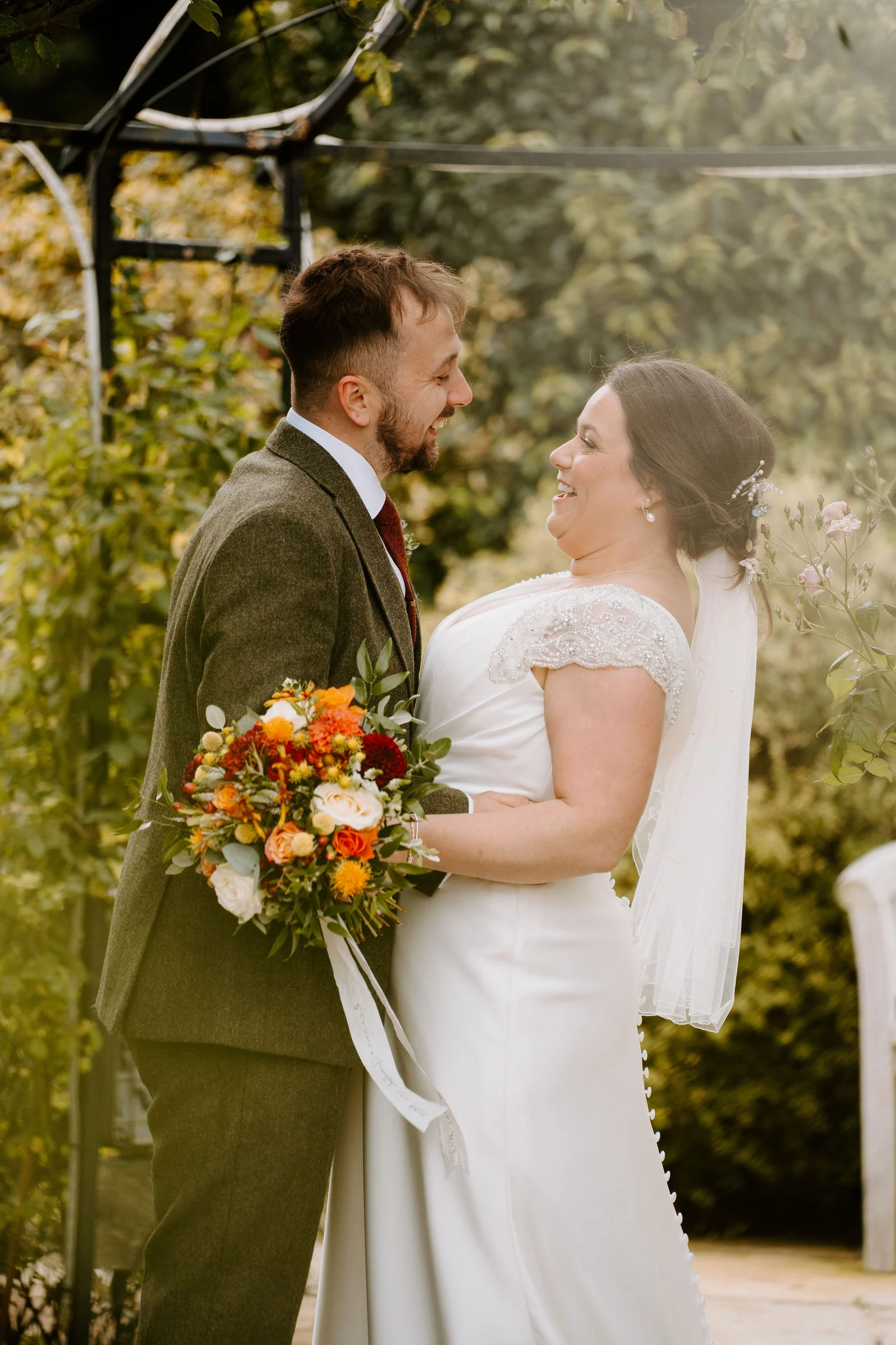 A groom in a green tweed suit and a bride in a white dress with a short veil smile at each other under a leafy garden arch, she holding an autumn bouquet of orange and red flowers.