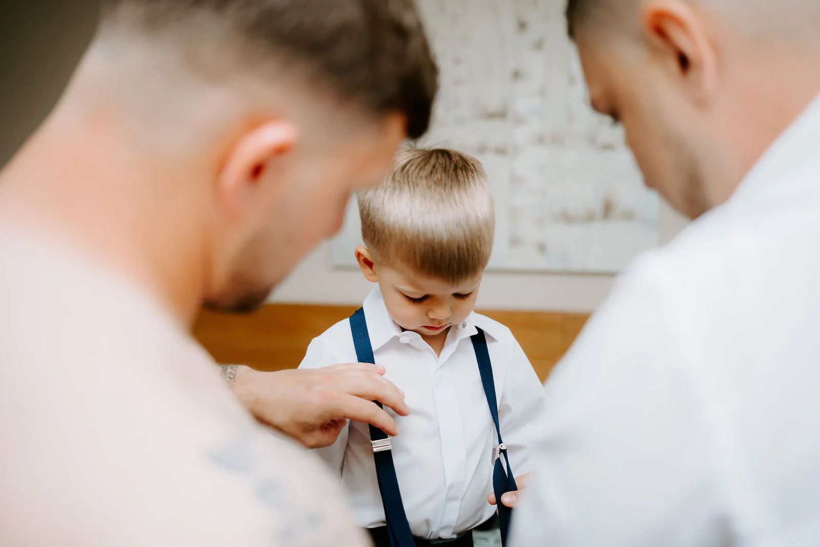 Two men fasten the navy braces on a small boy in a white shirt in a bright room before the ceremony