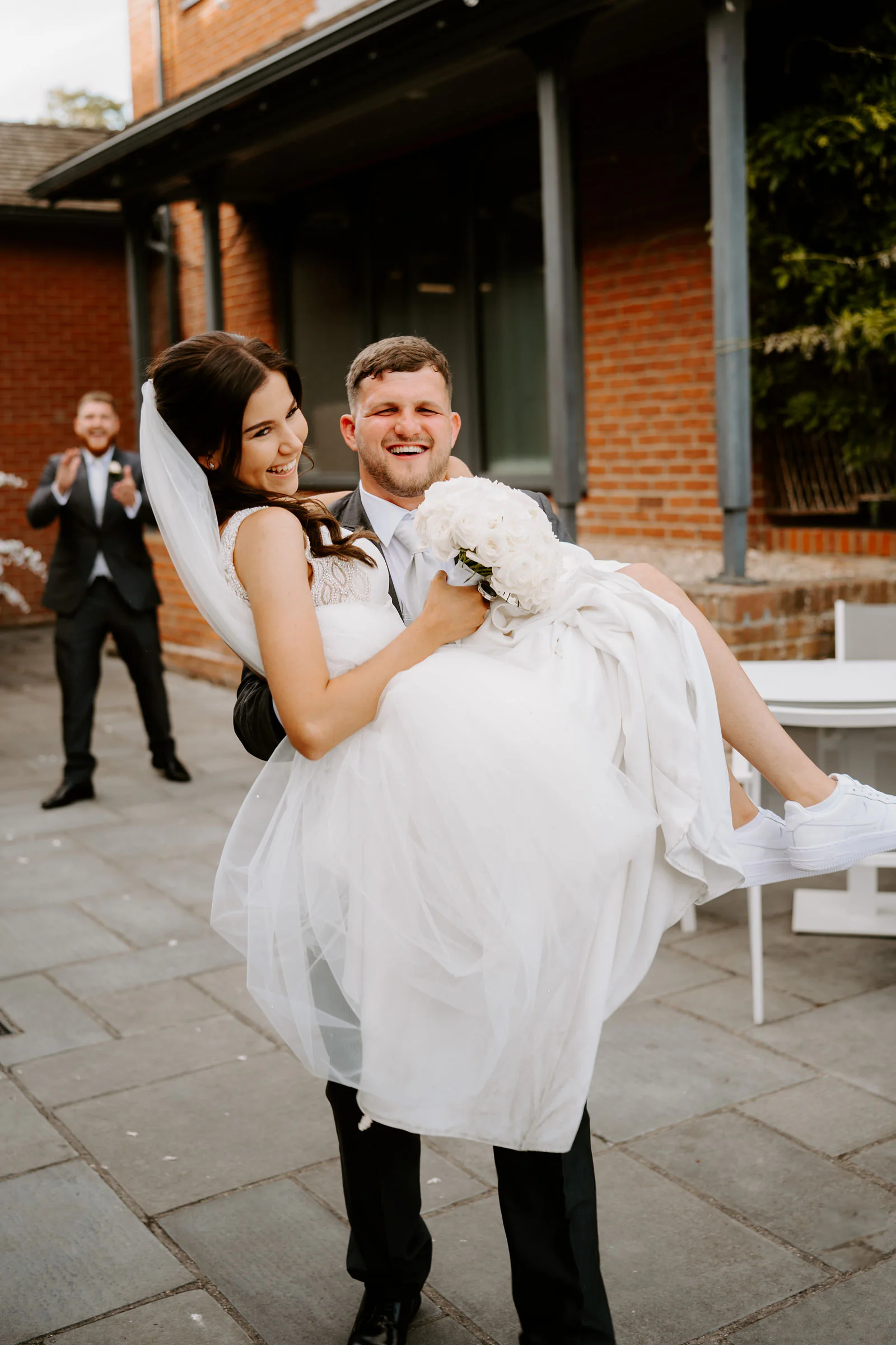 Jordan carries Molly in his arms across a paved terrace in front of a red-brick building while a friend cheers behind them