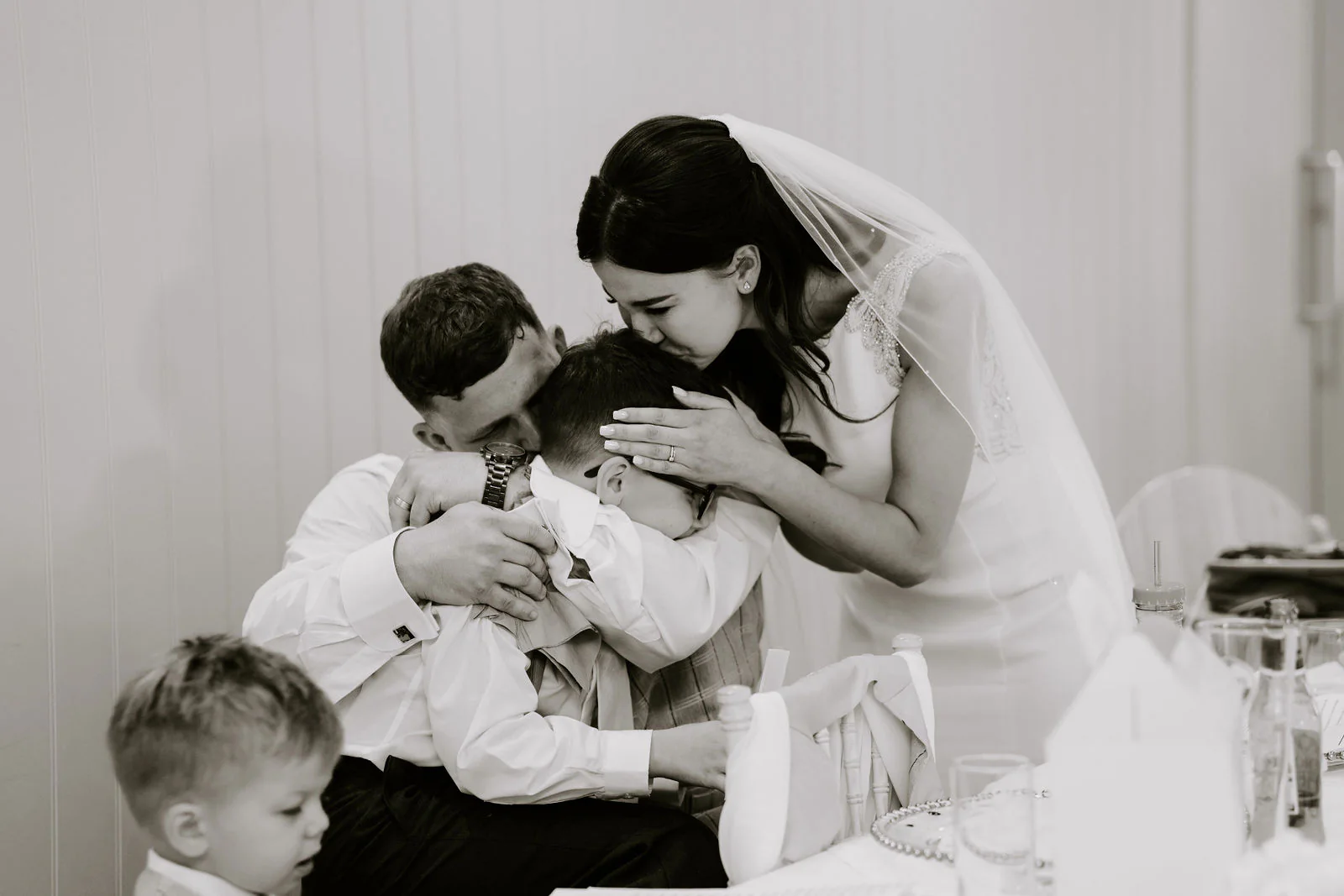 In black and white, Molly in her veil bends to hug a small boy and a man at the table while another boy looks on
