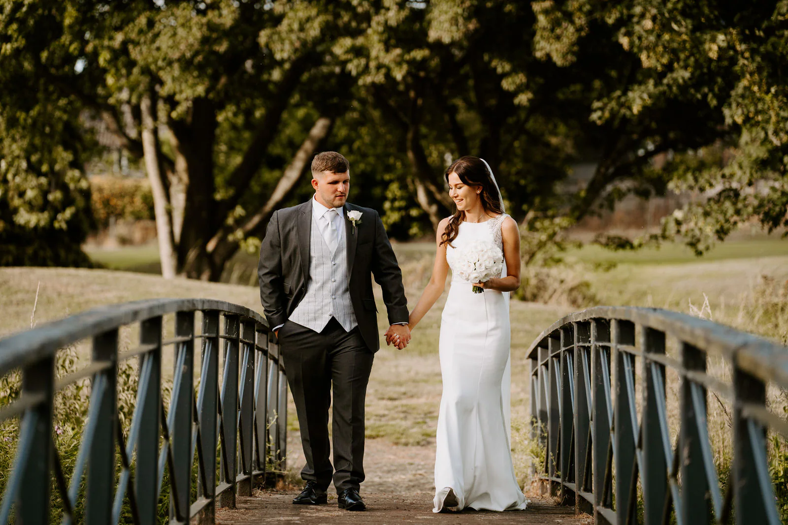 Jordan in a grey suit and Molly in a fitted white gown walk hand in hand across a small green footbridge with trees behind them