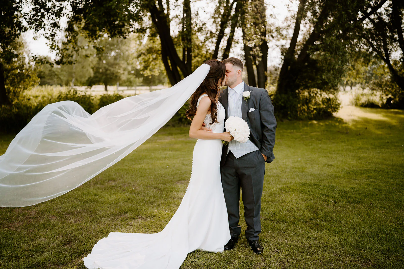 Molly and Jordan kiss on a lawn beneath tall trees as her long veil blows out behind her