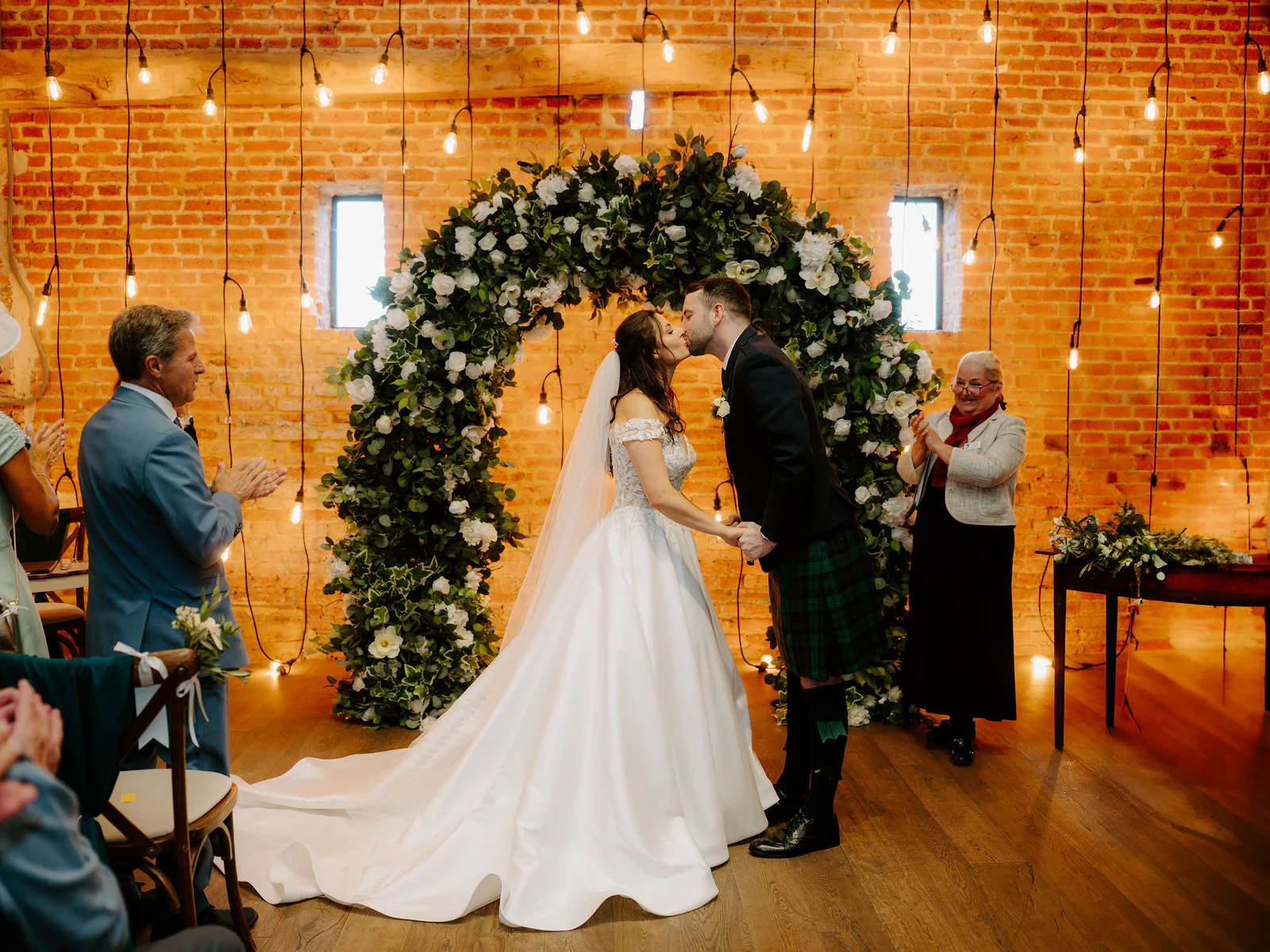Rose in a long veil and Ryan in a green tartan kilt kiss beneath a floral arch in front of a brick wall of festoon lights while two guests applaud