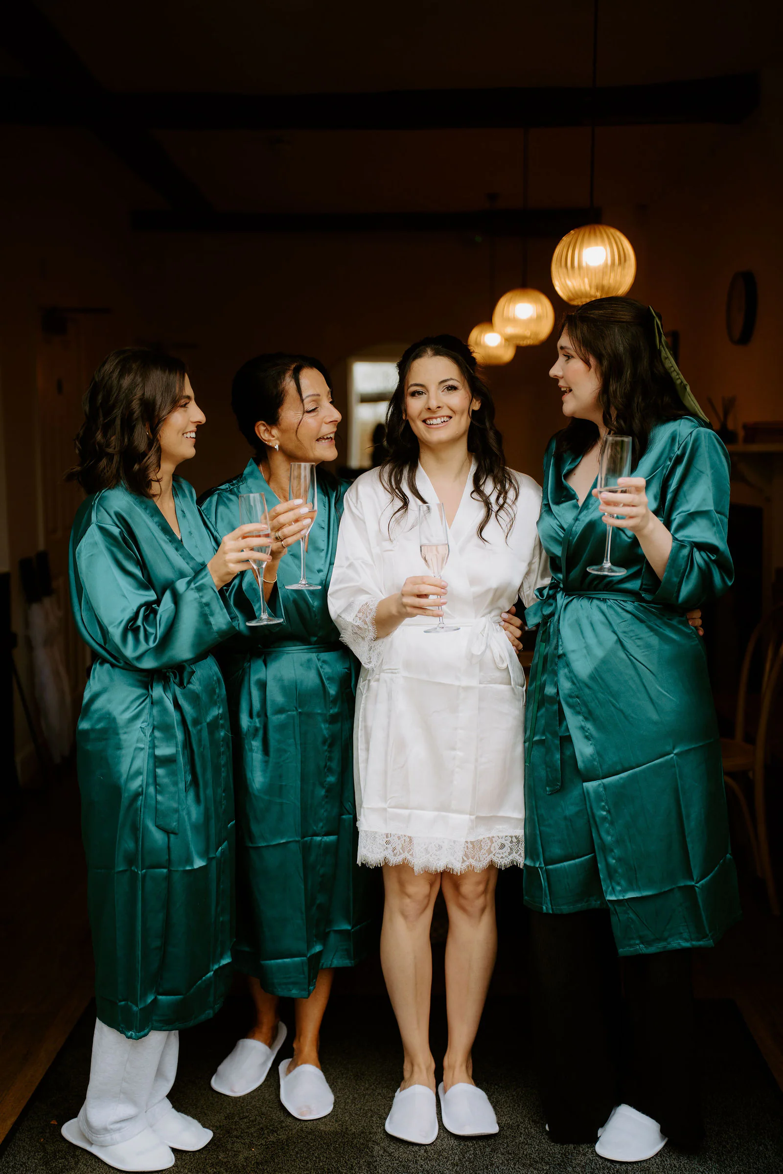Rose in a white robe stands laughing with three bridesmaids in teal satin robes, all holding glasses, in a dark room with warm pendant lights