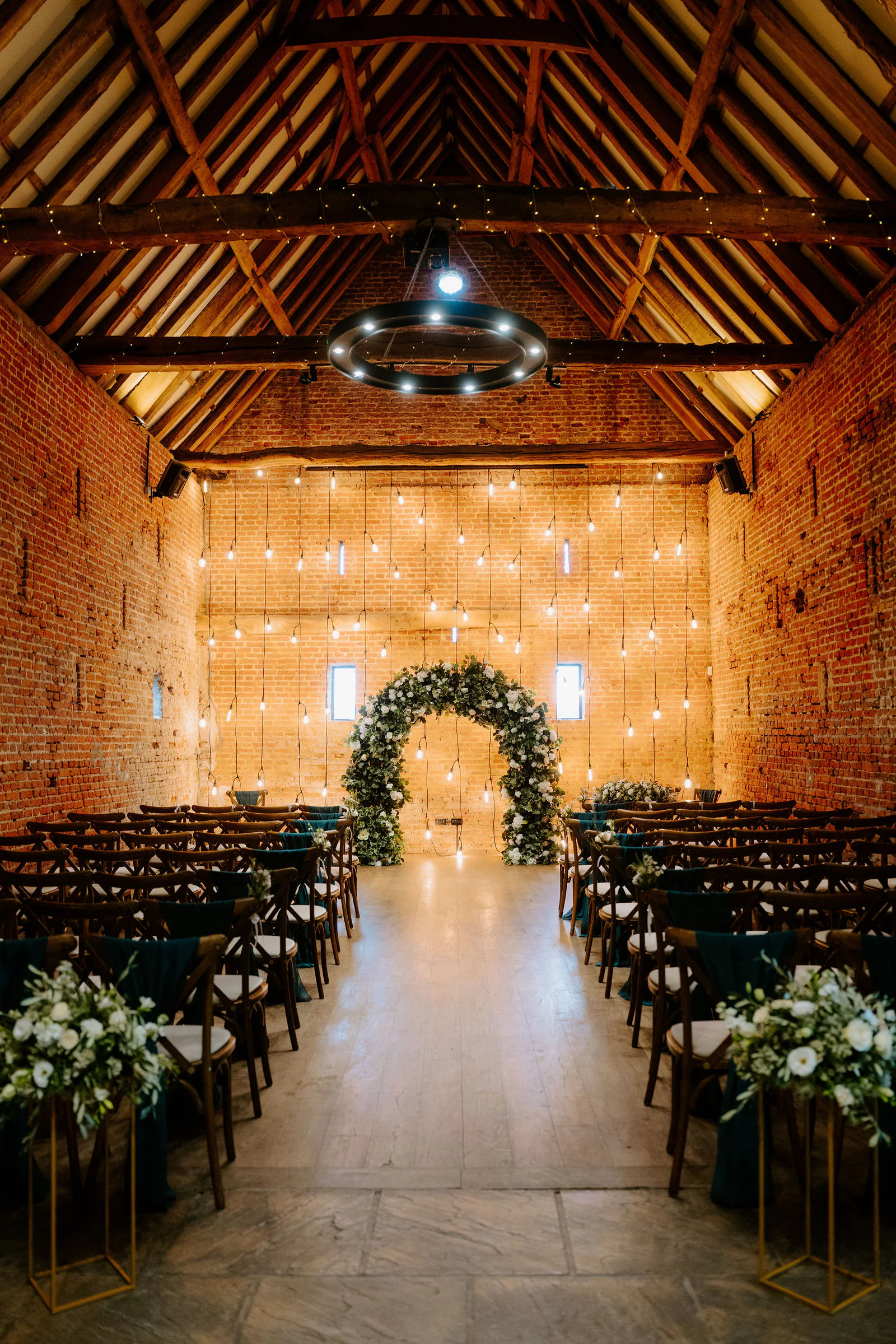 A brick barn with a high timber roof and walls of hanging festoon lights, set with rows of chairs facing a ring of white flowers and greenery