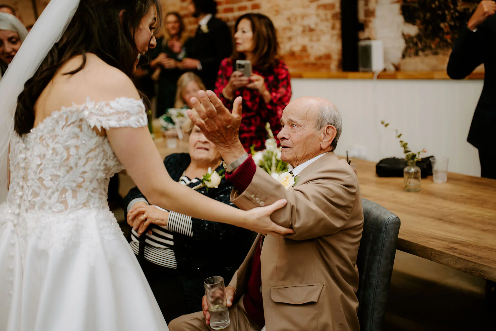 Rose in an off-the-shoulder lace gown reaches out to hold the hands of an older man in a tan jacket seated at a long wooden table