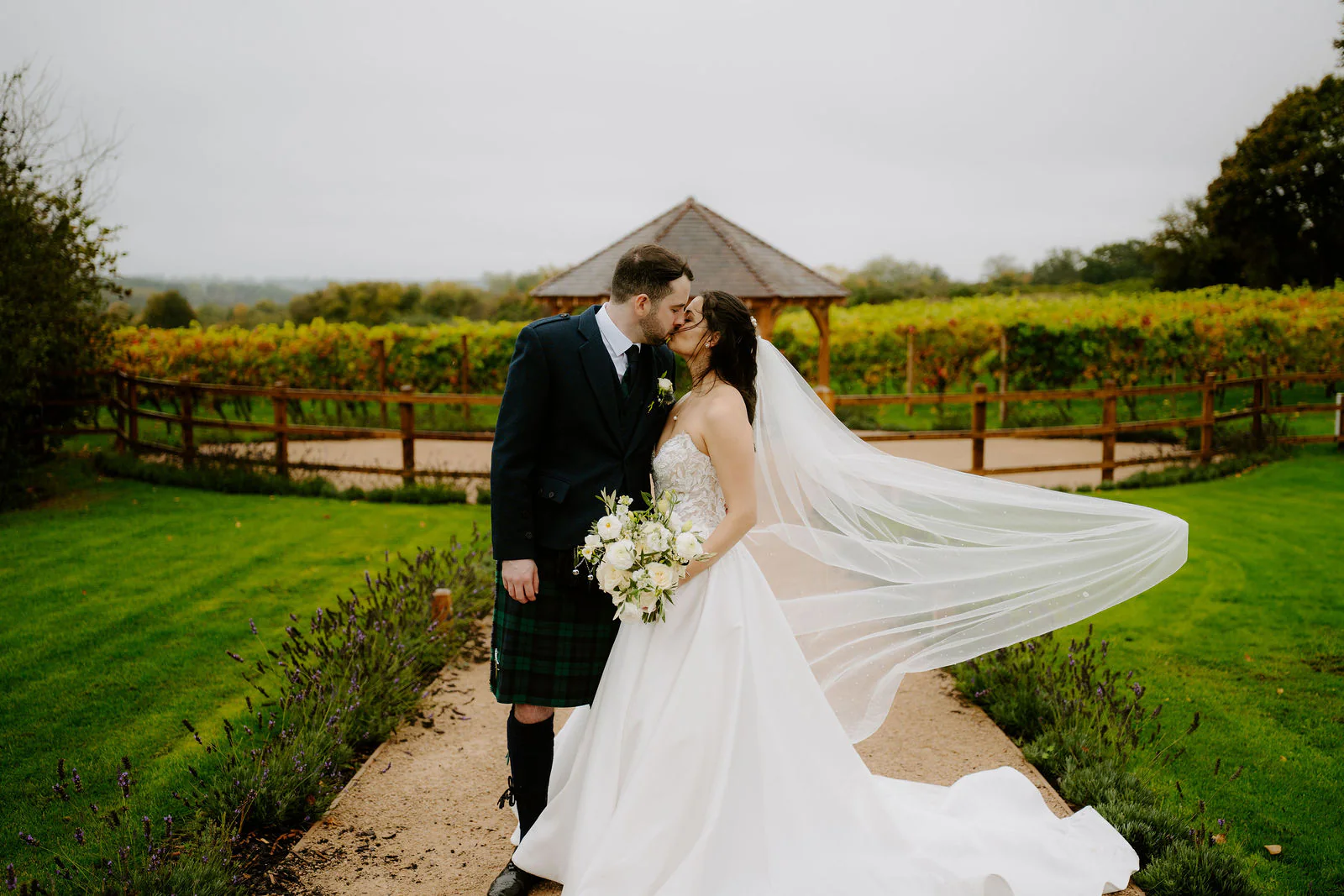 Rose and Ryan kiss on a gravel path between lavender beds with a vineyard and a timber gazebo behind them, Rose's long veil streaming in the wind
