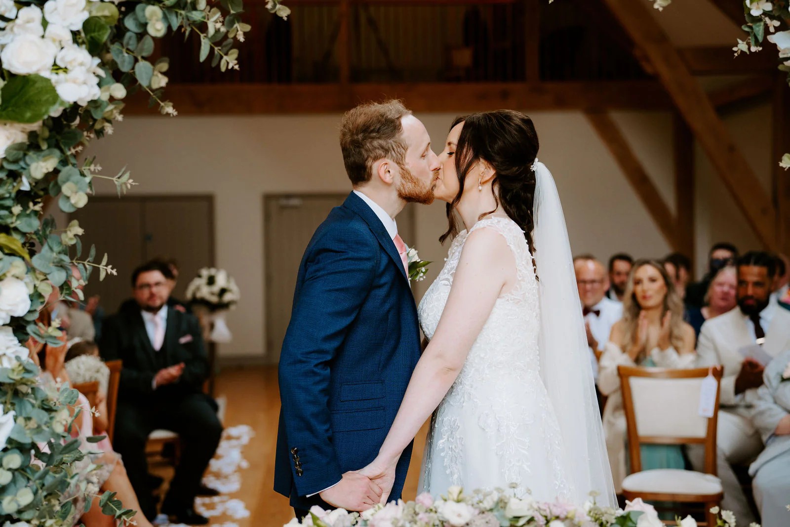 Sophie and Daniel kiss during their ceremony in a timber barn beside a tall arrangement of white flowers and greenery, guests seated behind them