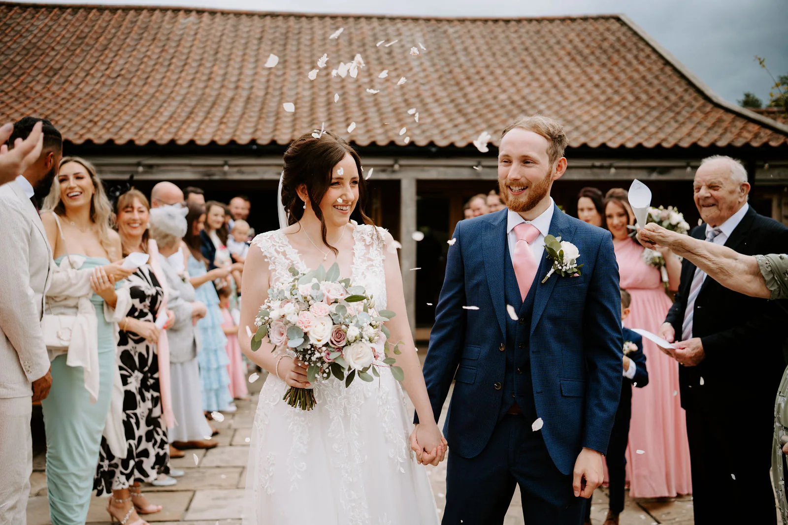 Sophie and Daniel walk hand in hand through thrown petals between friends and family beneath a clay-tiled barn roof at Easton Grange