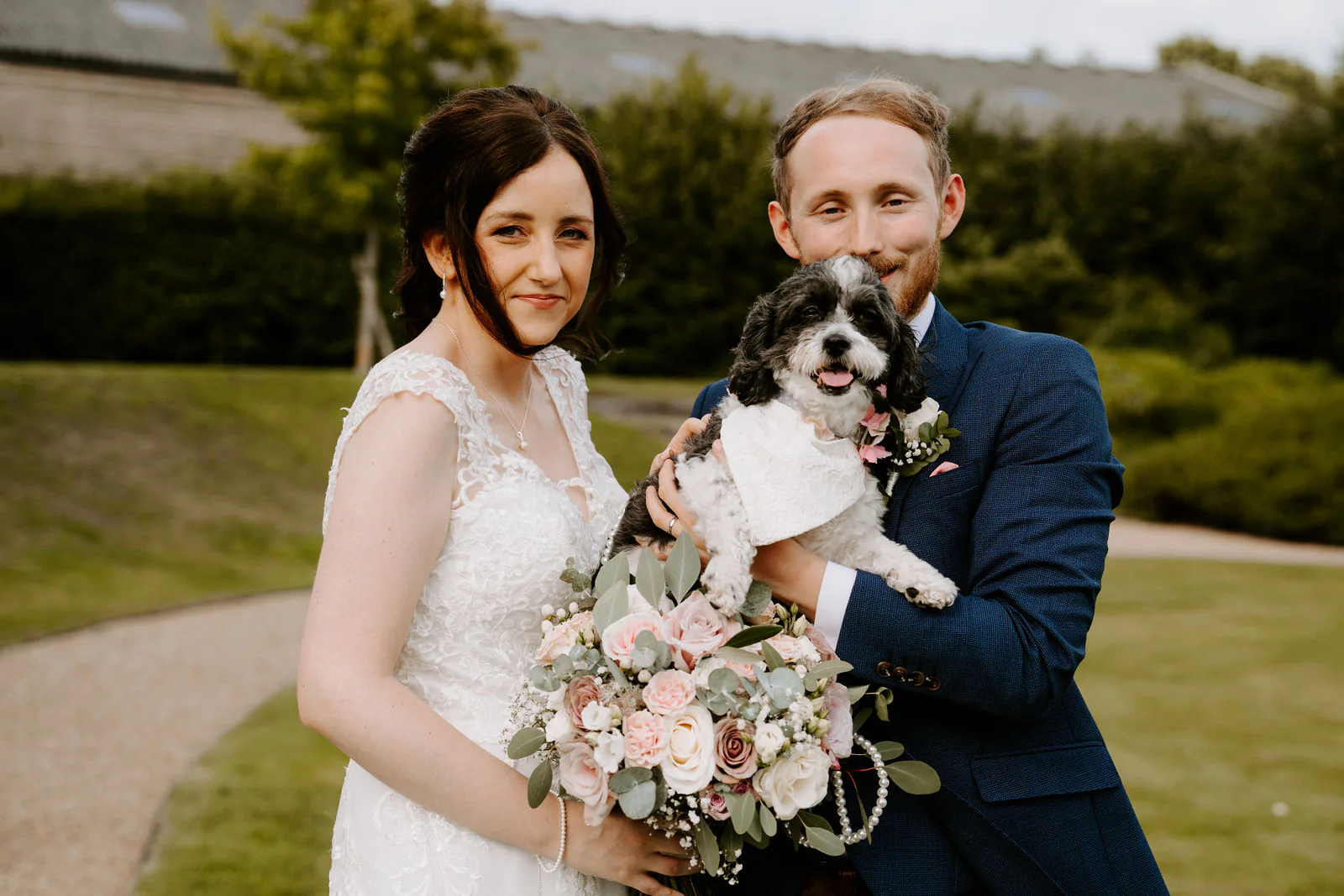 Daniel holds the couple's small black and white dog in a bow while Sophie holds a bouquet of blush roses on the lawn at Easton Grange