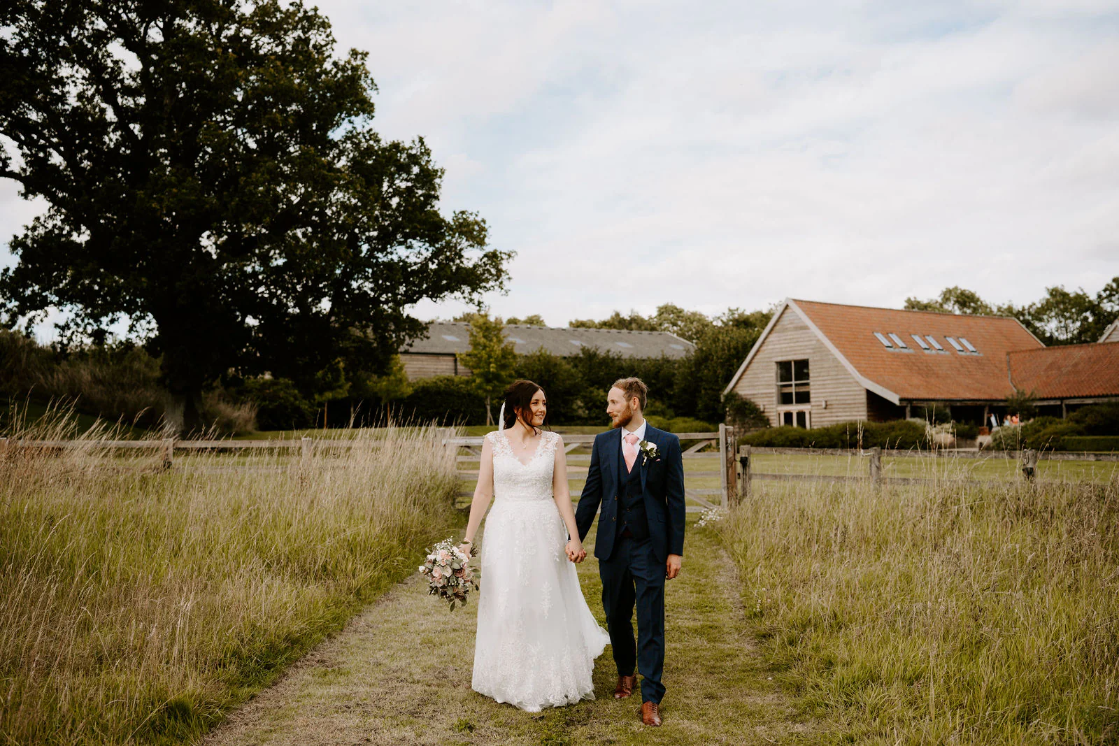 Sophie and Daniel walk hand in hand along a mown path through long grass towards a timber-clad barn with a tiled roof
