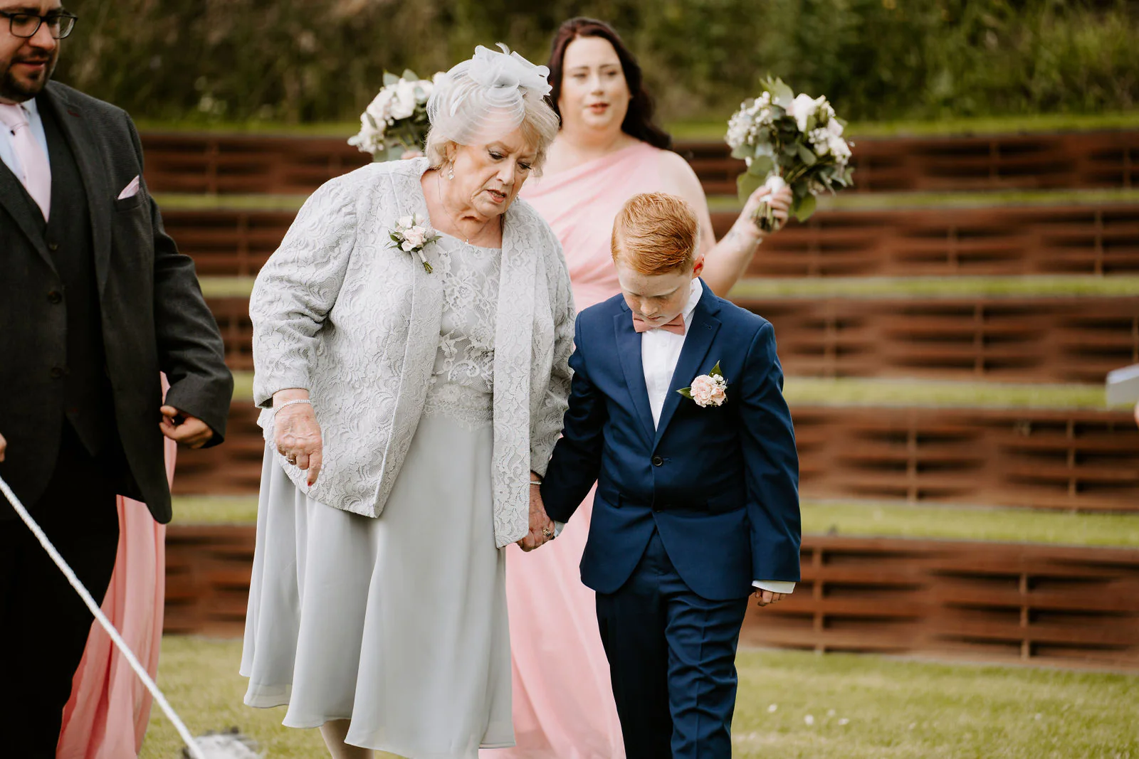 An older guest with a corsage walks hand in hand with a boy in a navy suit beside terraced walls of rust-coloured steel