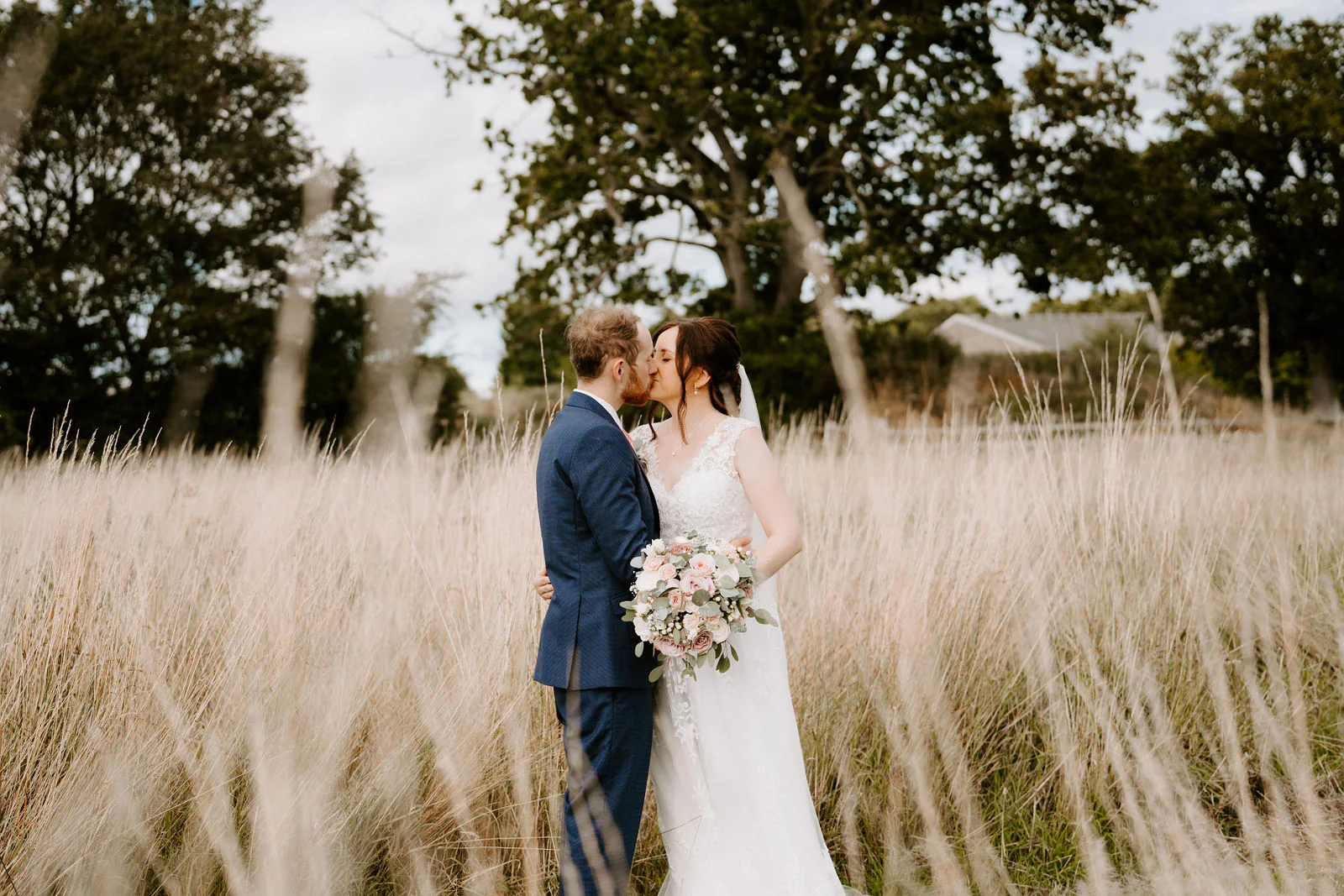 Sophie and Daniel kiss in tall pale grass with trees behind them, Sophie holding a bouquet of blush roses