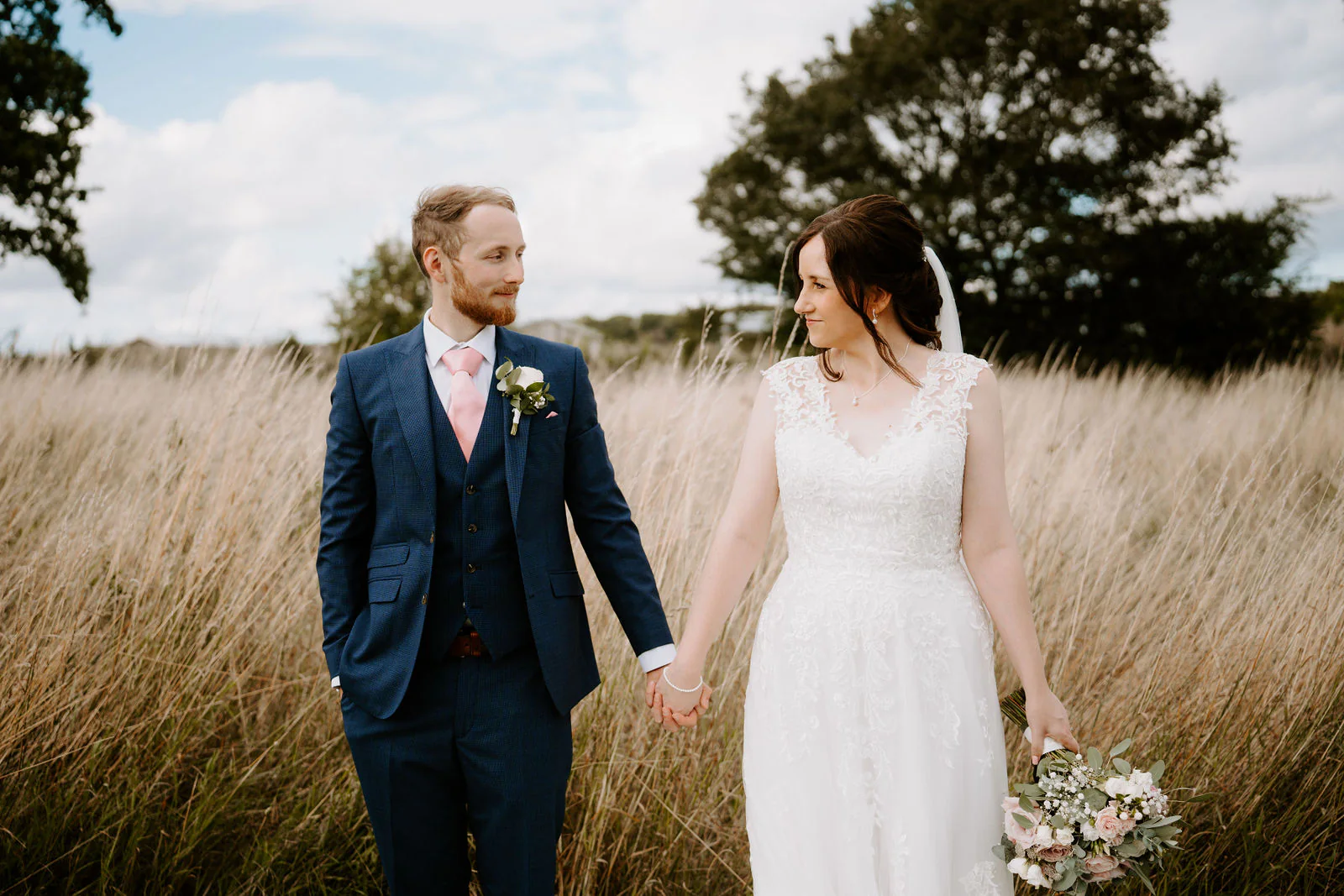Daniel in a navy suit and Sophie in a lace gown hold hands and look at each other in tall pale grass beneath large trees