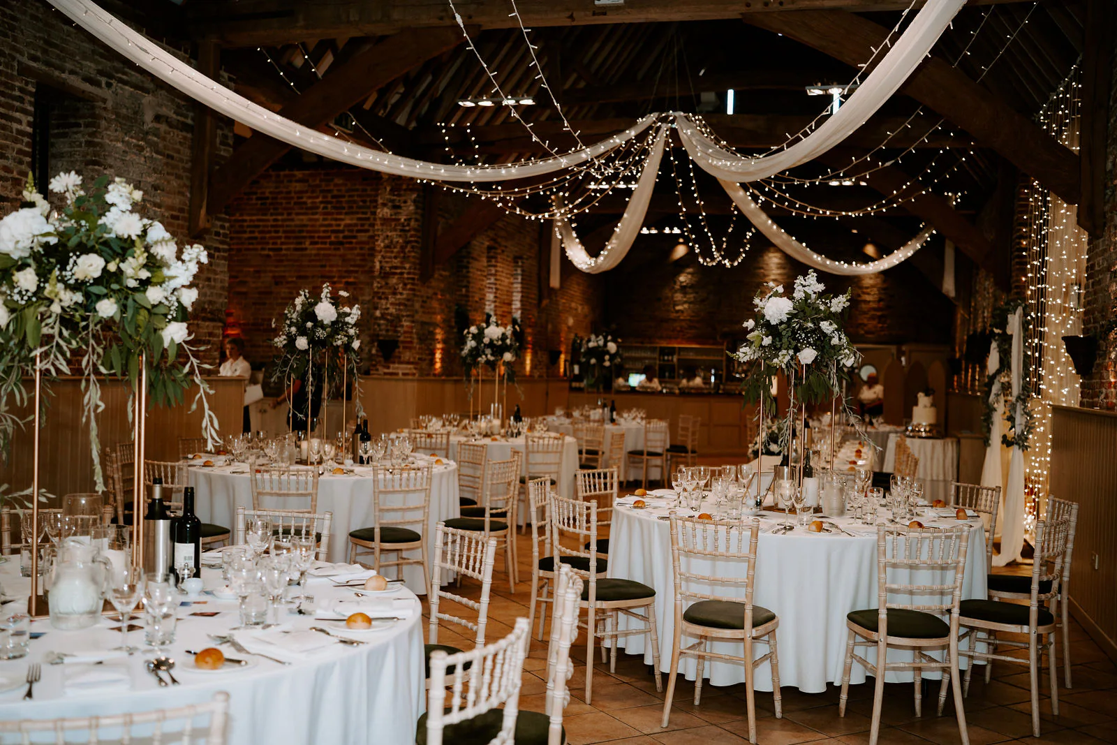 Round tables laid with white linen and tall flowers beneath draped fabric and strings of lights in a timbered brick barn