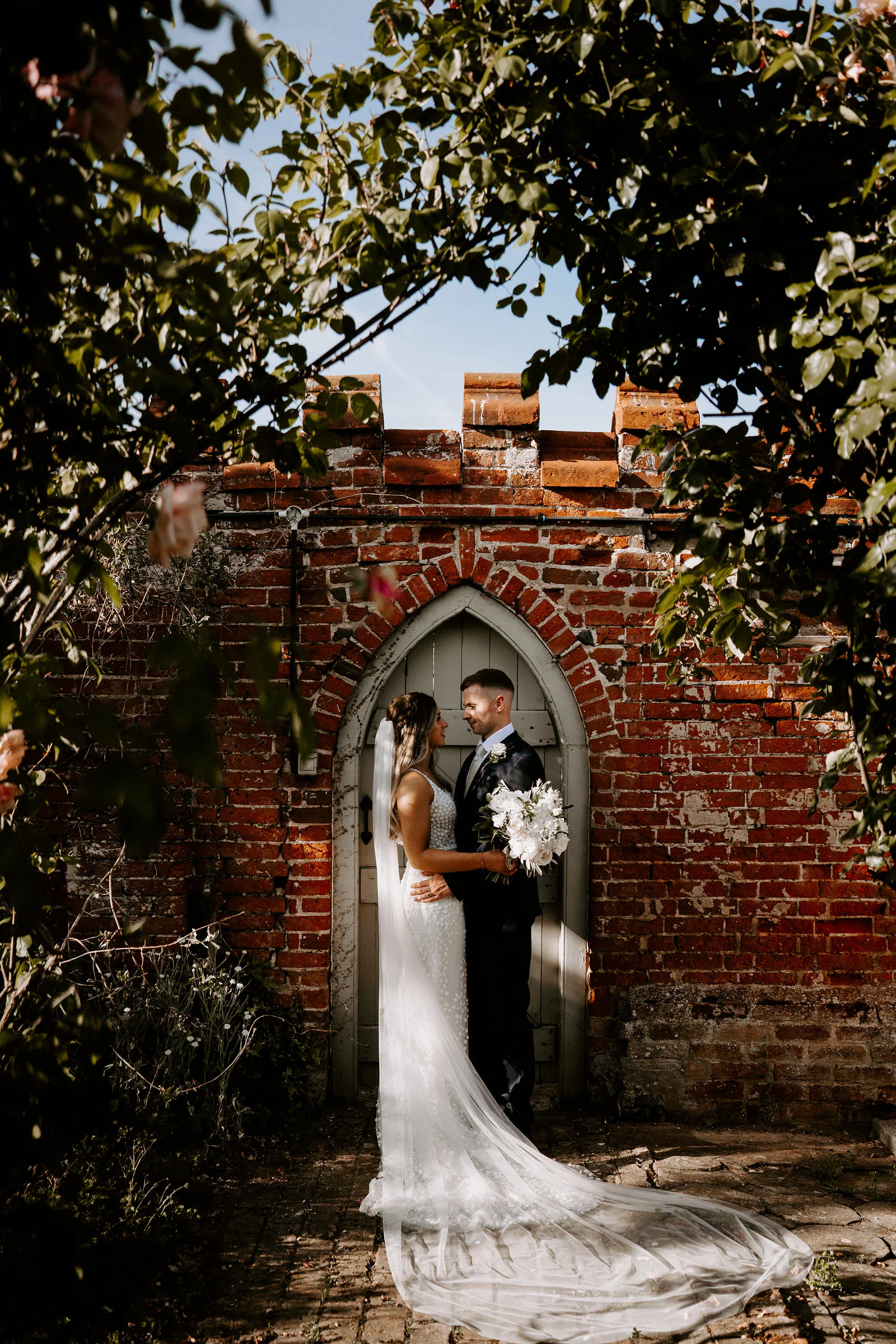 Shantelle in a long lace gown and Karl in a dark suit stand in a pointed doorway in a red-brick garden wall with a battlemented top, framed by leaves